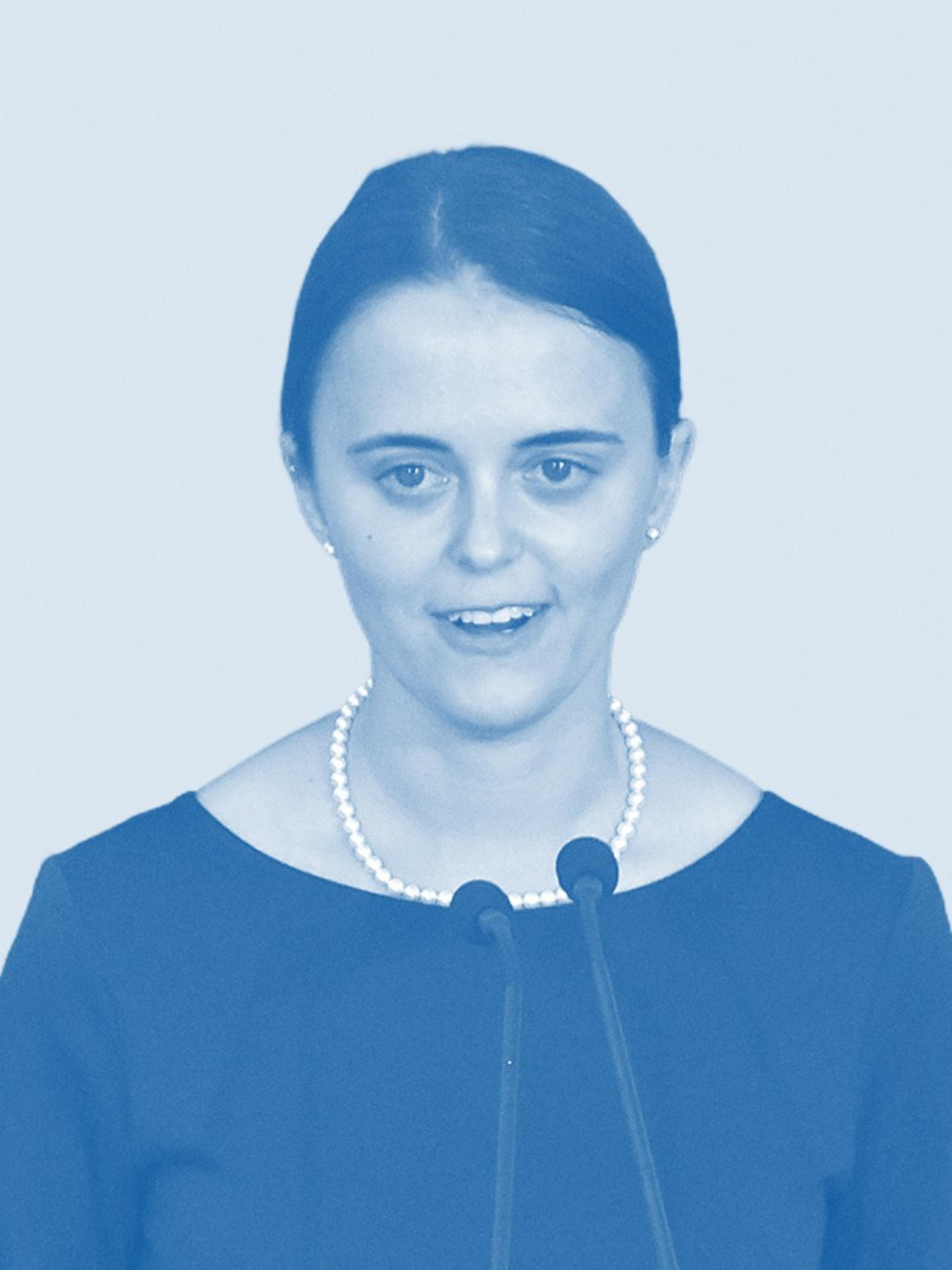 A woman with tied-back hair, wearing a dark top and a pearl necklace, is speaking at a microphone. The image has a blue tint.