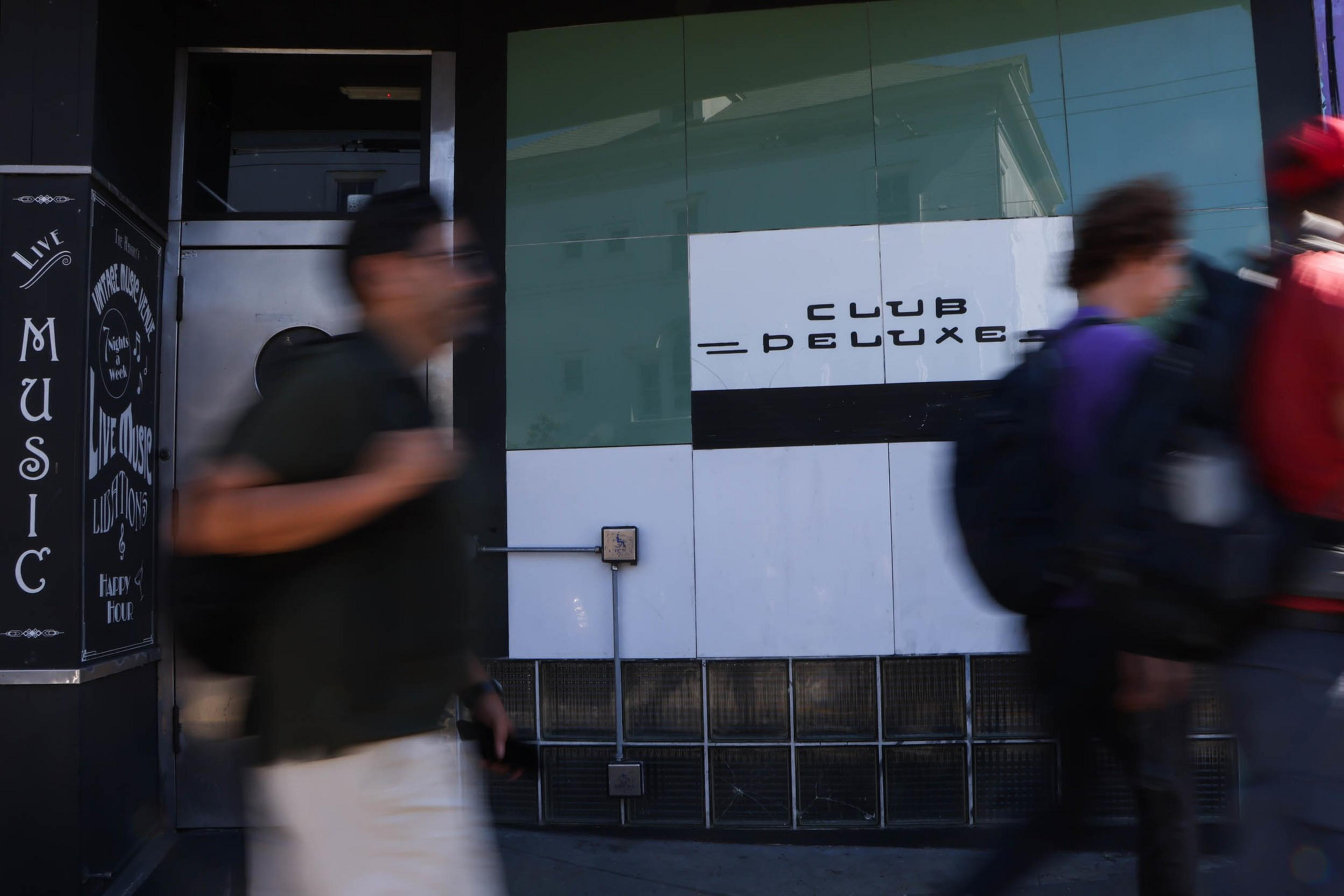 A club entrance with a sign reading "Club Deluxe" above glass blocks, and three blurred people walking past, one carrying a backpack.