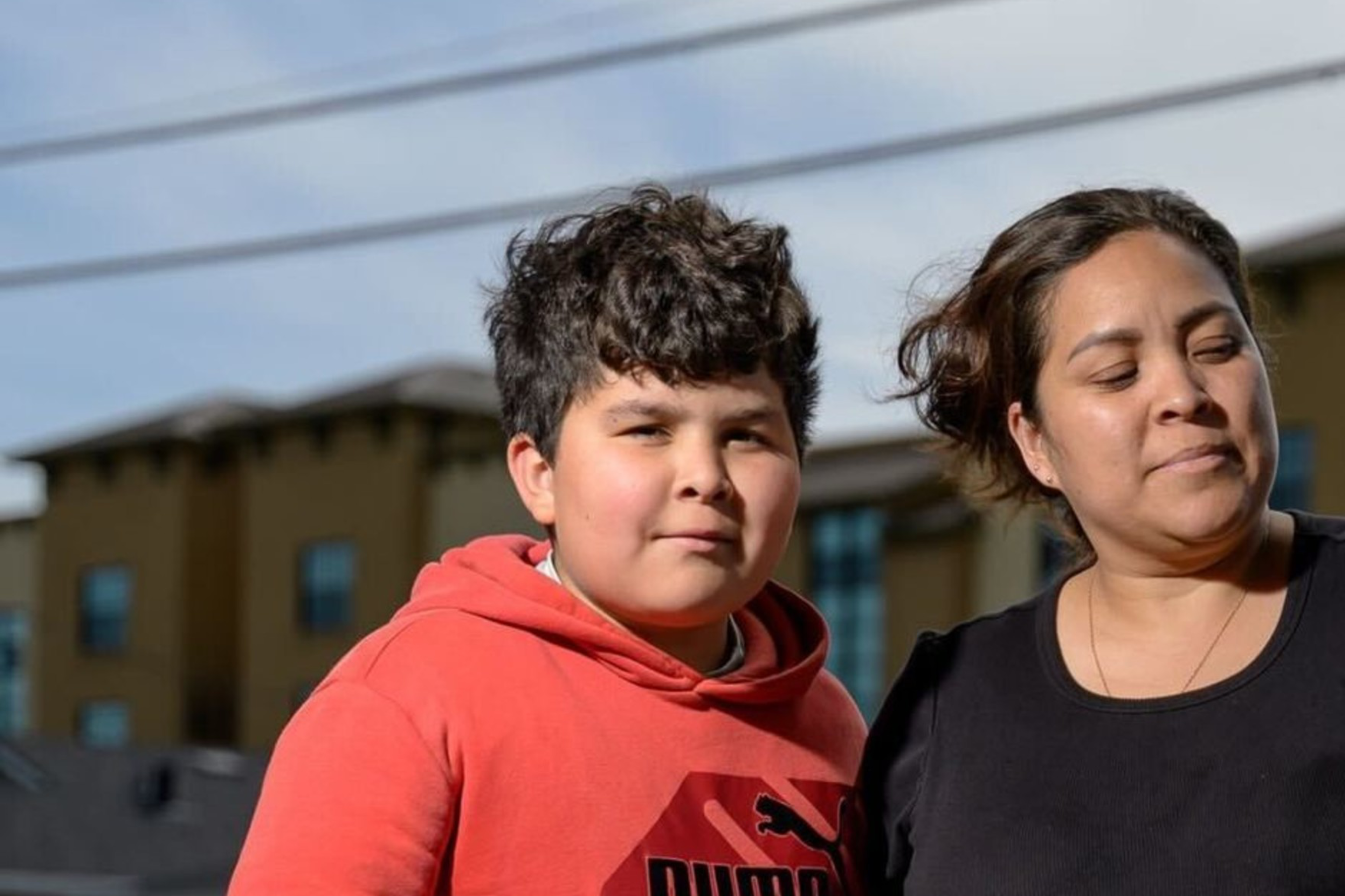 A woman stands on a sidewalk with her arms around two boys. One wears a red hoodie; the other has a backpack. They are in front of a building under a cloudy sky.