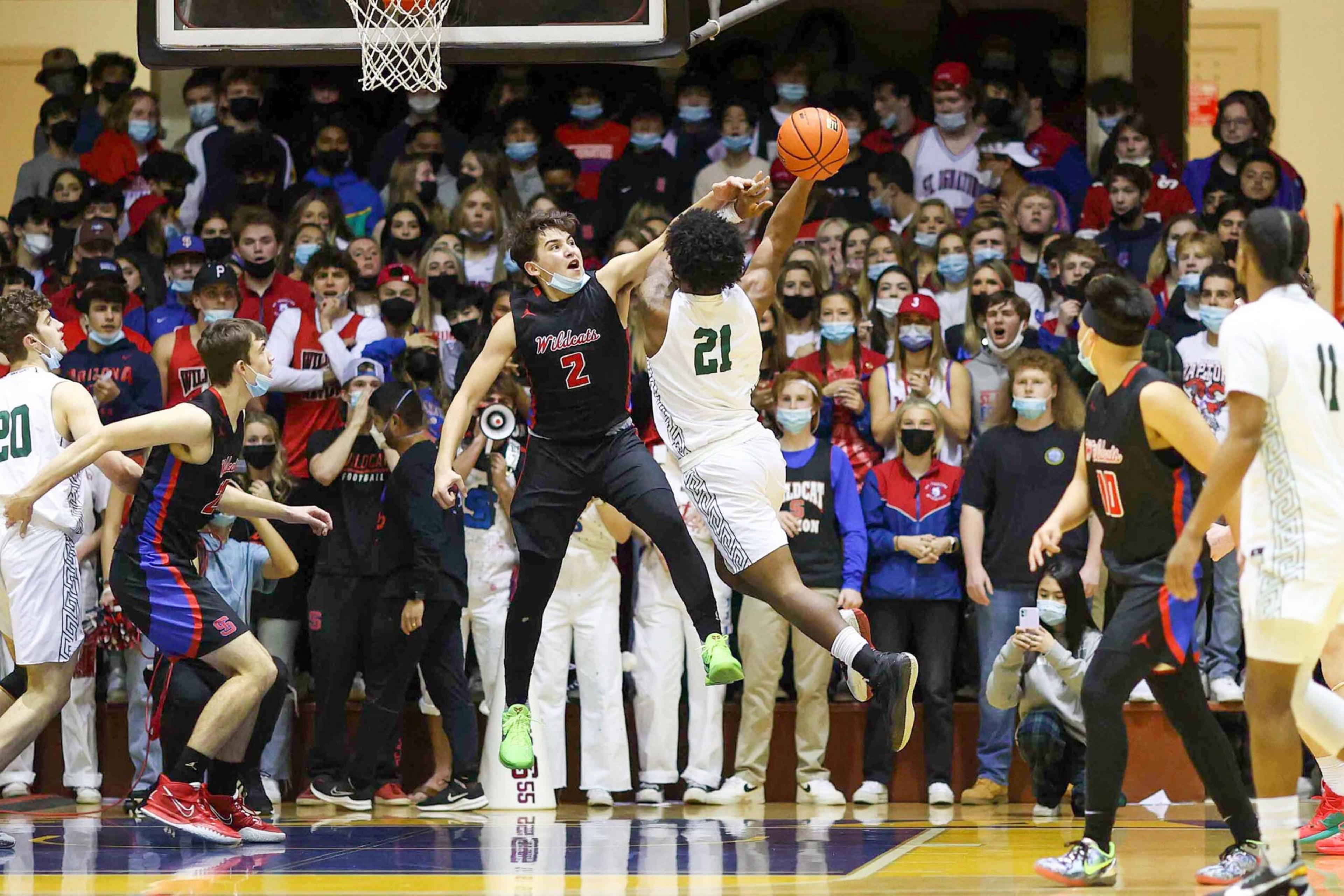 A basketball player in white jersey 21 jumps for a shot while a player in black jersey 2 tries to block him amid a crowded gym with cheering fans.