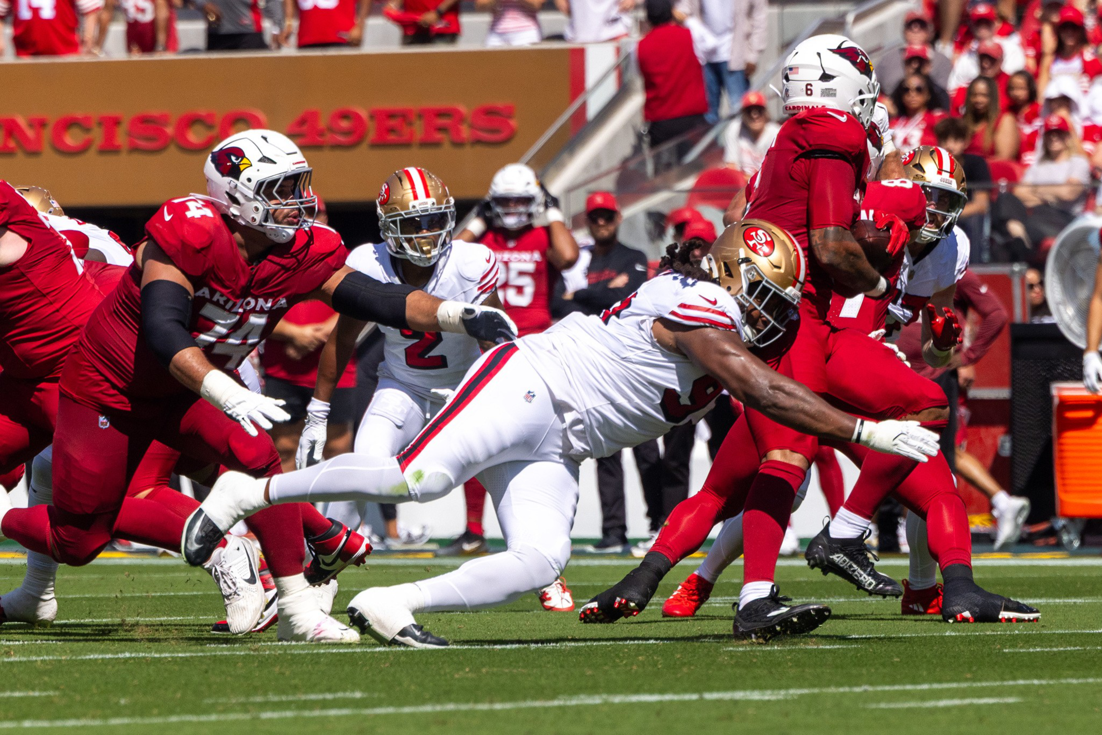 A player in white dives to tackle a player in red holding the football, while other players from both teams run nearby on a football field.