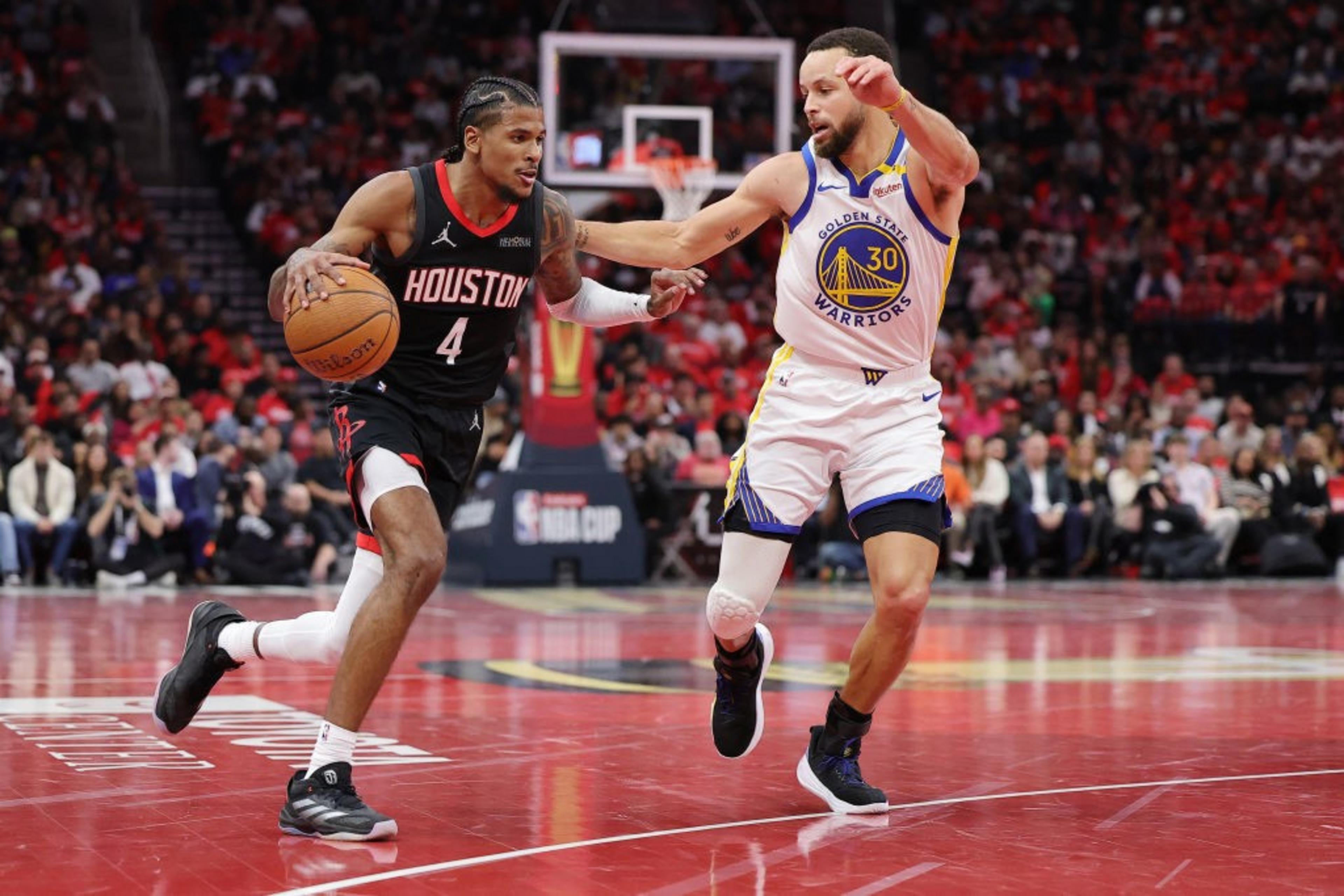 Two basketball players are on the court. One in a Houston jersey dribbles the ball while the other, in a Golden State jersey, defends him. The crowd watches intently.