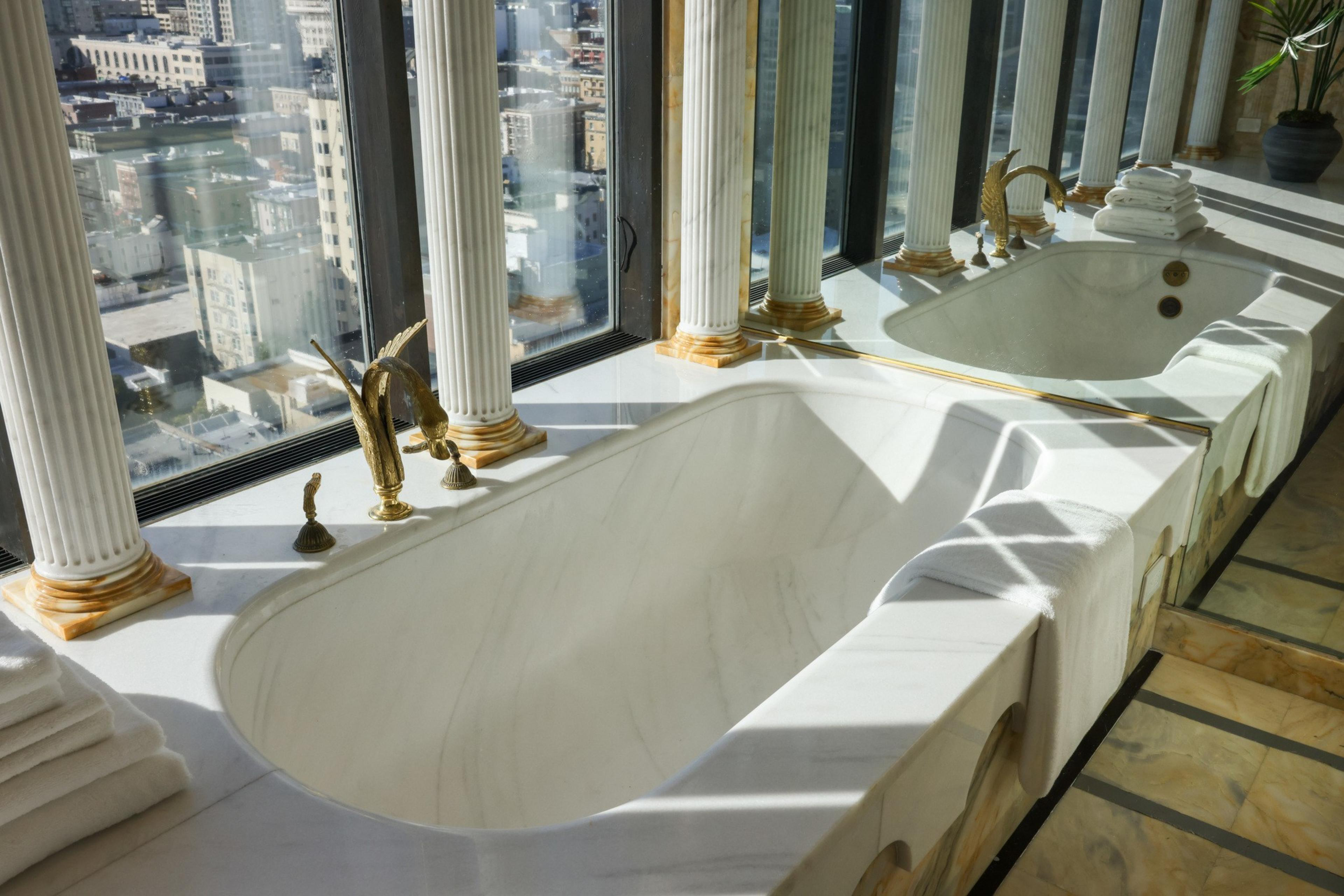 A luxurious marble bathtub with elegant gold fixtures is set beneath tall windows offering a city view. Tall white columns and neatly folded towels complete the scene.