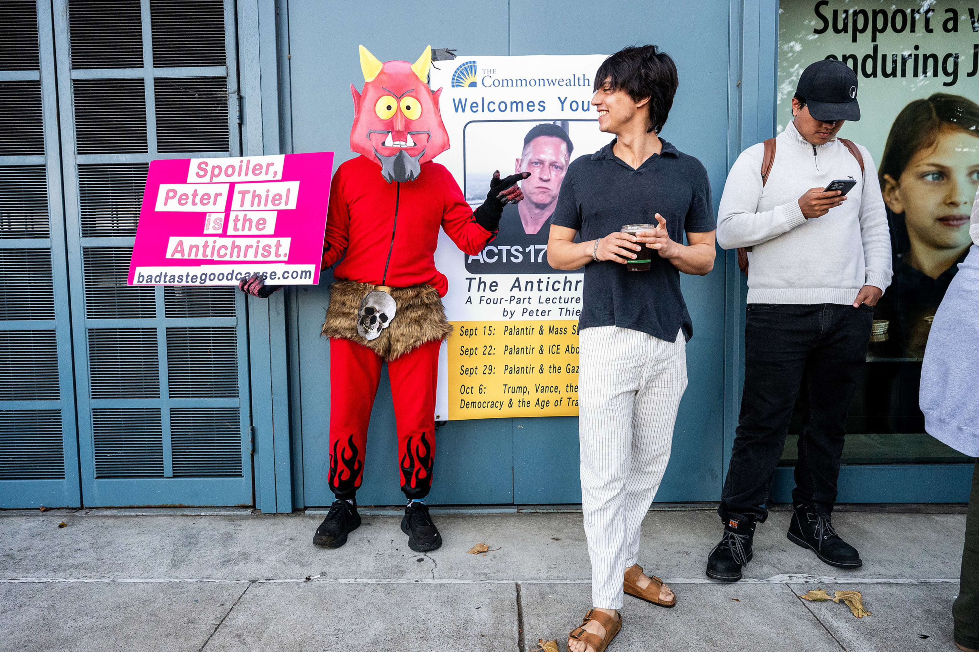 A person in a red devil costume holds a sign calling Peter Thiel the Antichrist while two casually dressed people stand nearby, one smiling.