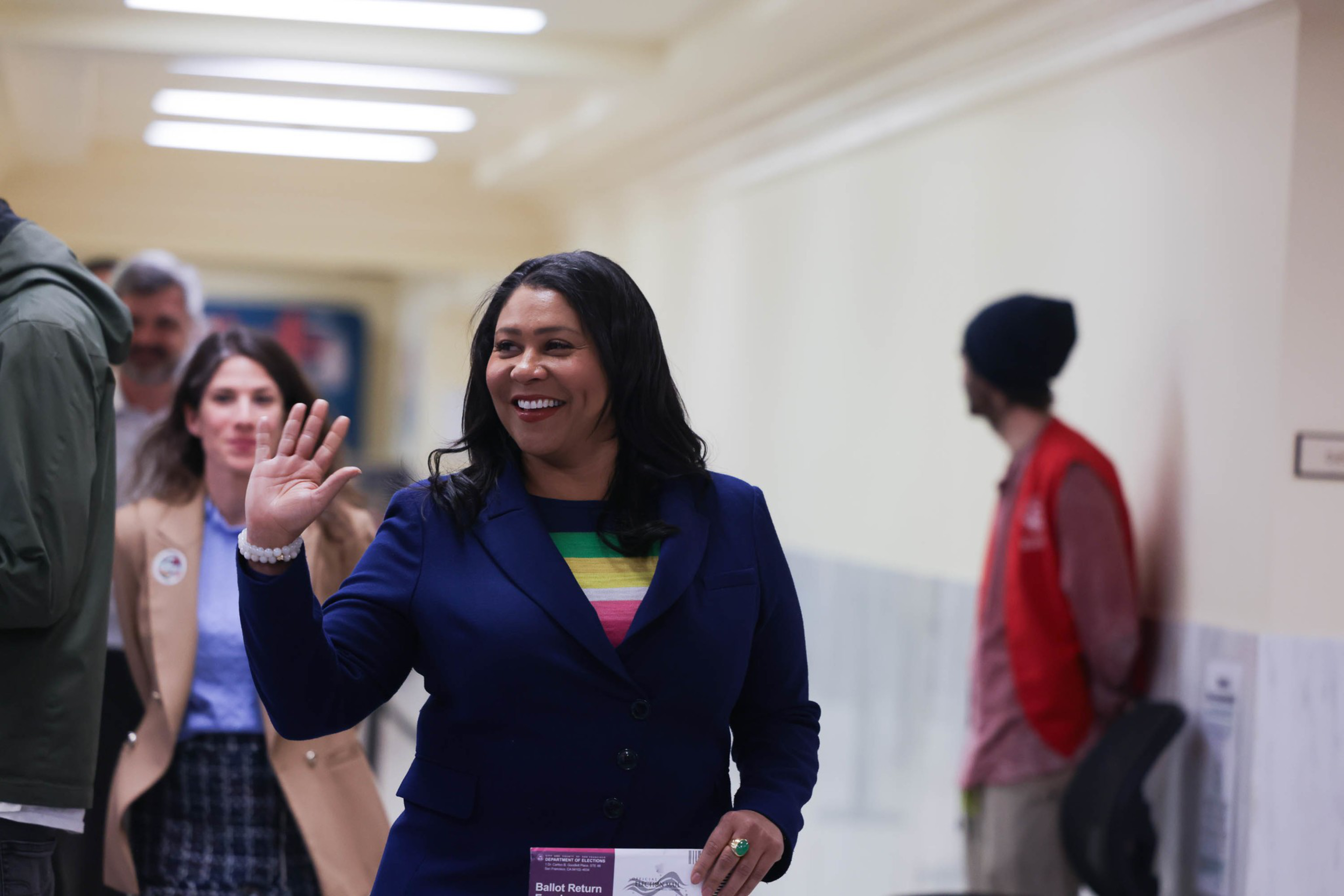 A smiling woman in a blue blazer waves as she walks through a crowded hallway.