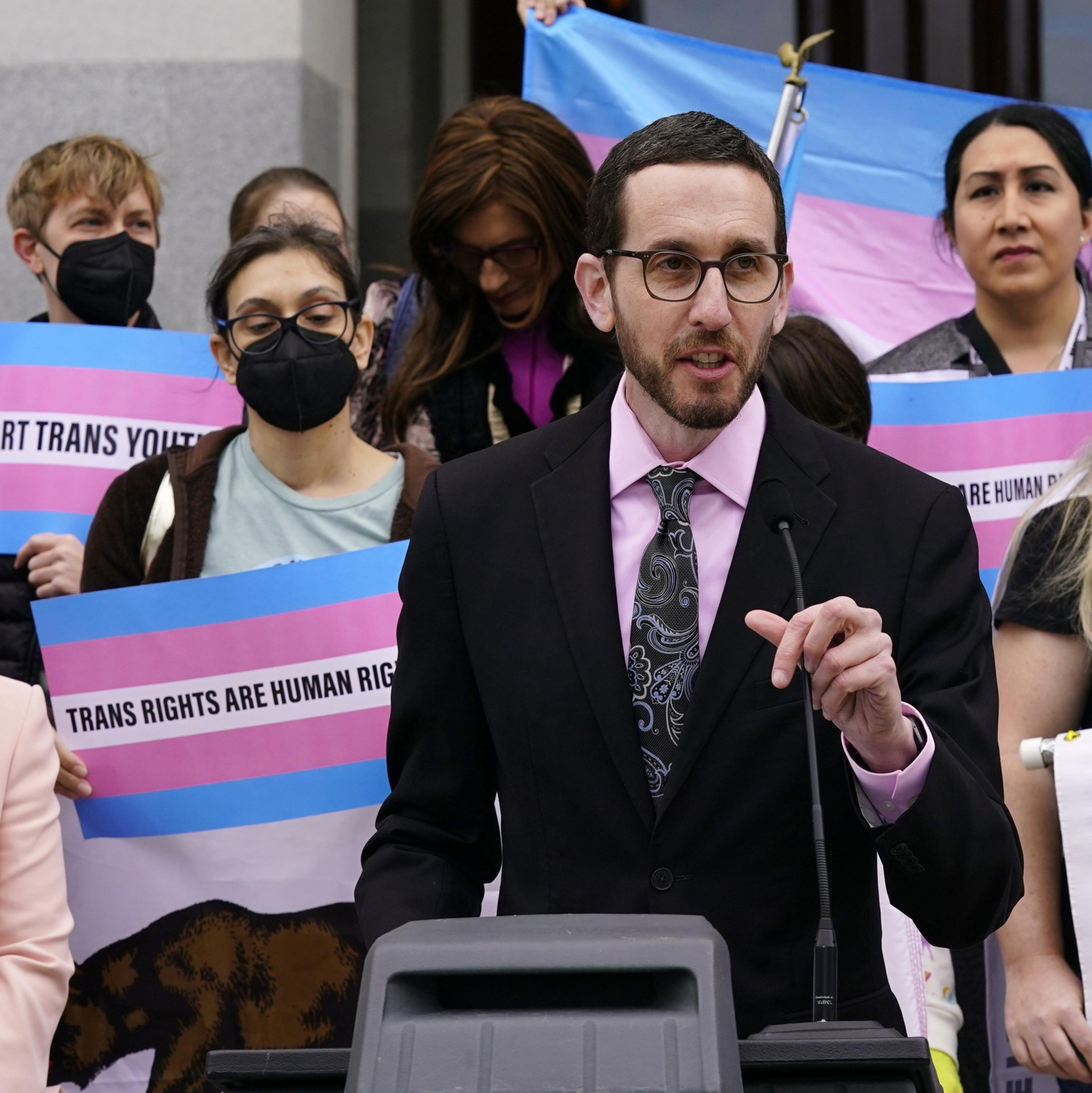 A person in a suit speaks at a podium with a crowd behind them holding pink and blue signs reading "Trans Rights Are Human Rights."