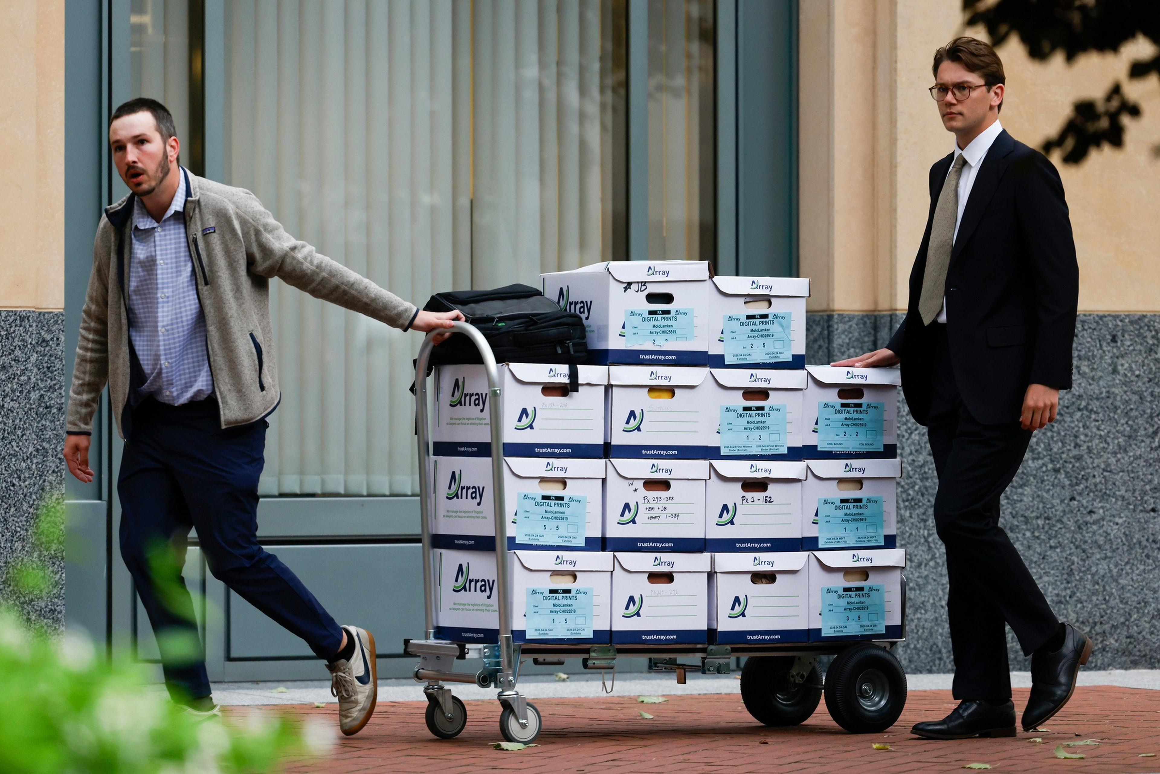 Two men, one in casual clothes and the other in a suit, move a cart stacked with white boxes labeled “Array” outside a building with large windows.