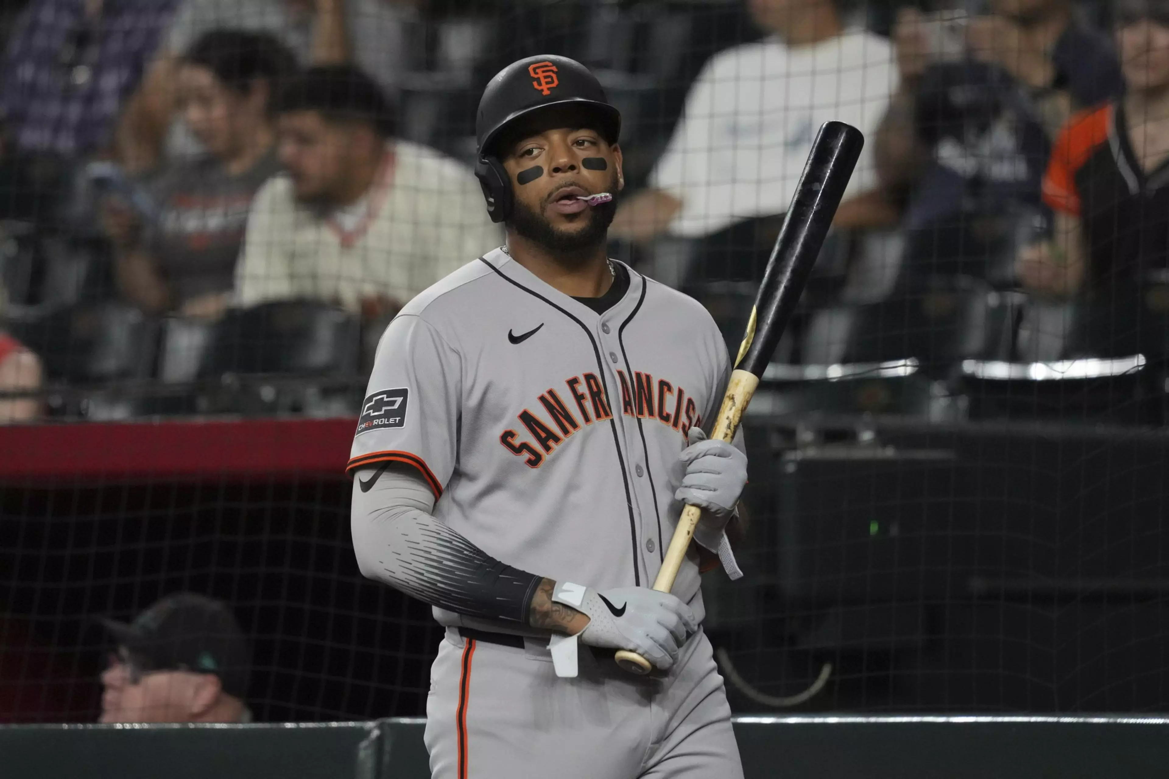 A baseball player in a gray San Francisco jersey and helmet holds a bat, wearing eye black and gloves, with fans seated in the background.