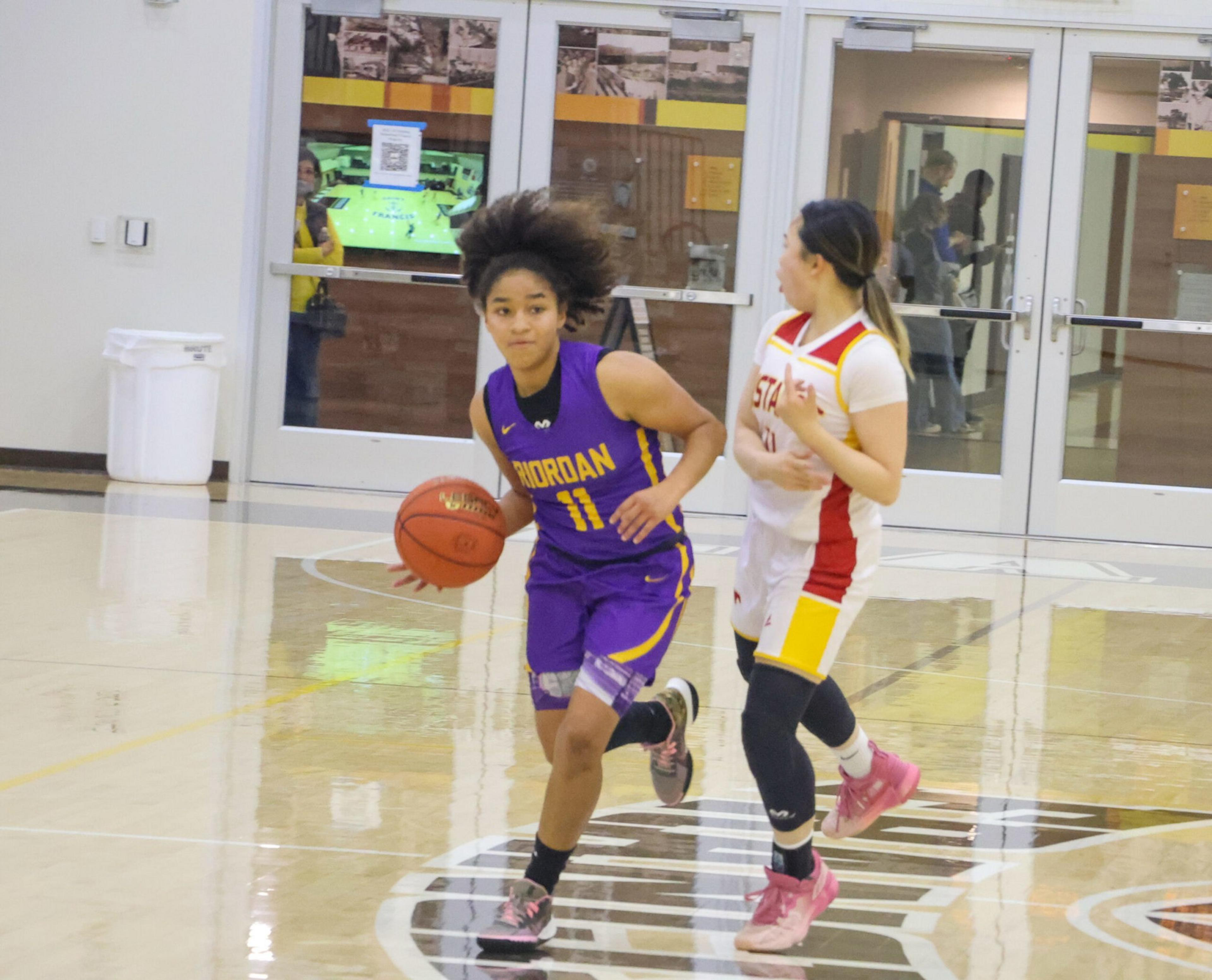 A basketball player in a purple jersey with "Jordan" and number 11 dribbles the ball while being closely guarded by a player in a white and red uniform.