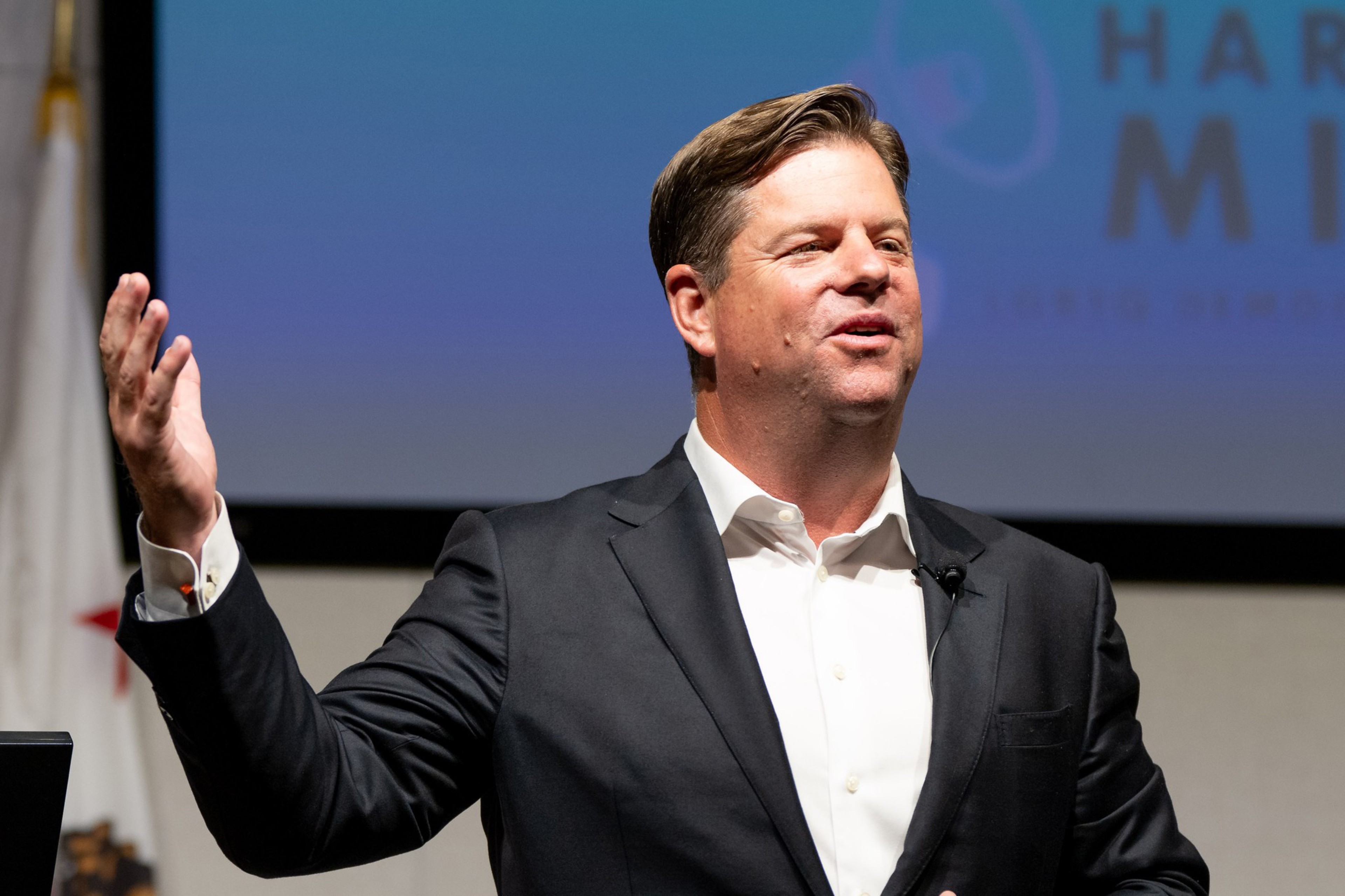 A man in a dark suit and white shirt is giving a speech, gesturing with one hand up. A flag is seen in the background, and a presentation slide is partially visible.
