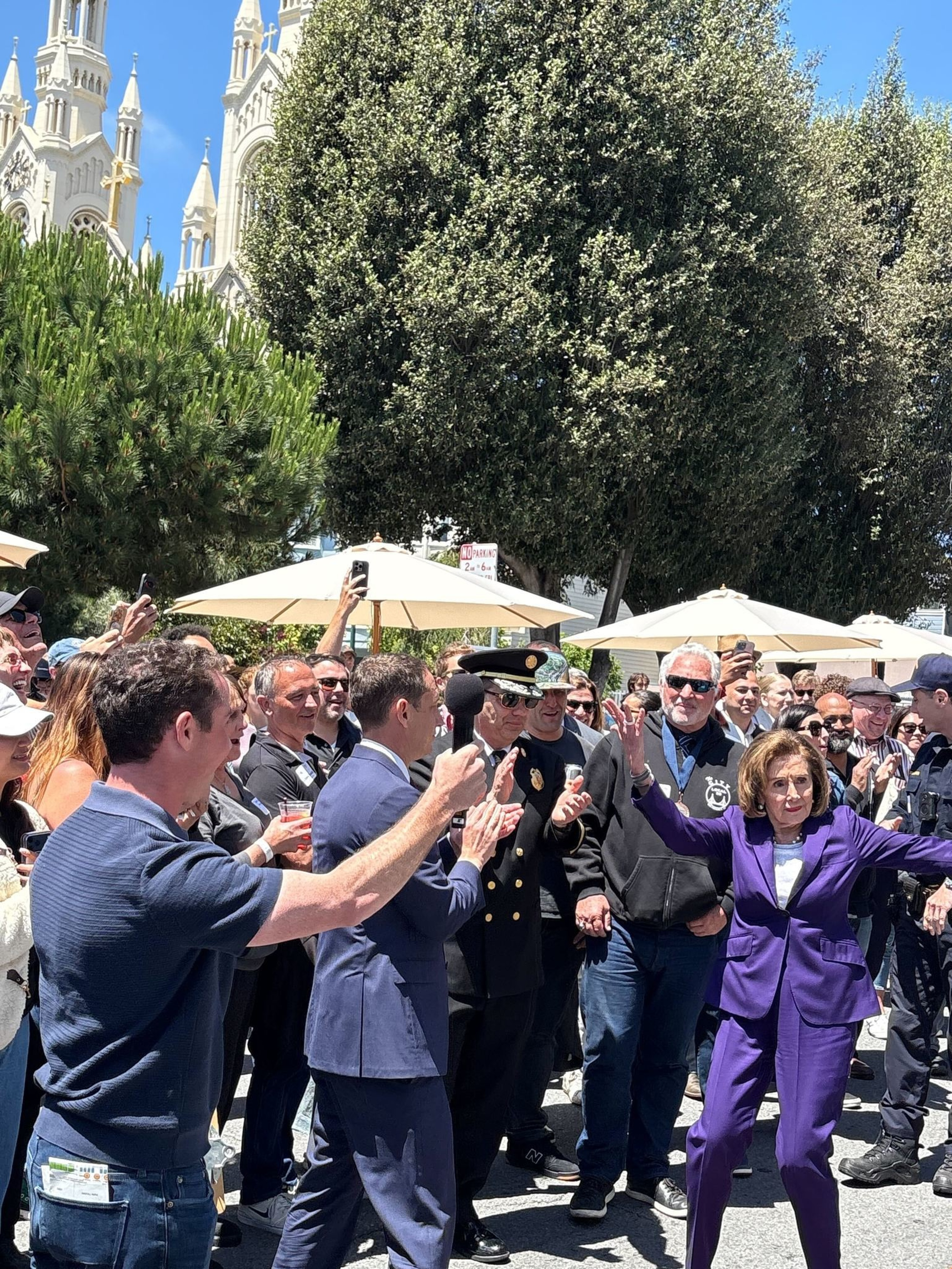 A woman in a purple suit gestures energetically while a crowd around her claps and cheers under sunny skies near large trees and a white church.