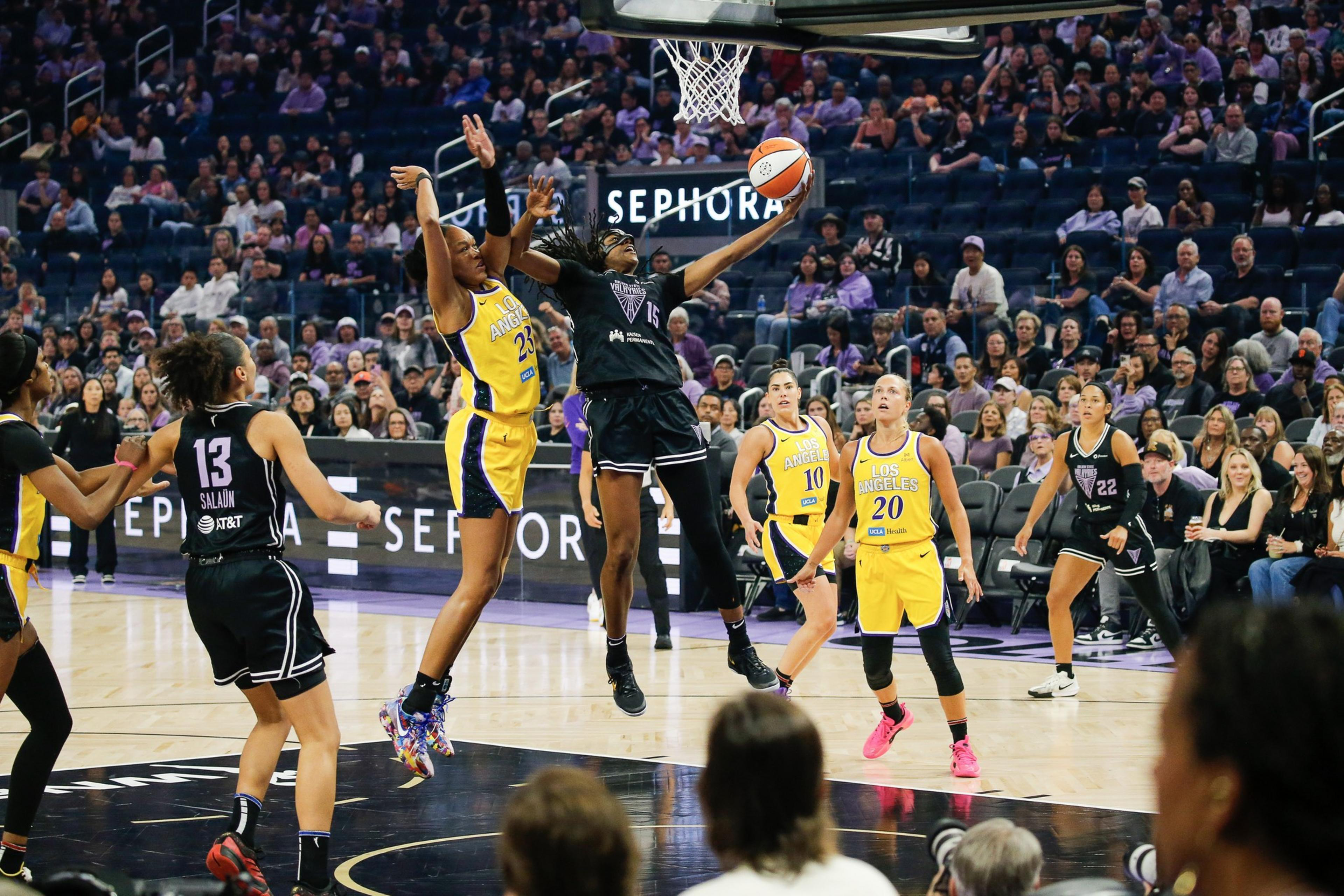 A basketball player in black jumps for a shot while a defender in yellow tries to block, with teammates and spectators watching intensely.