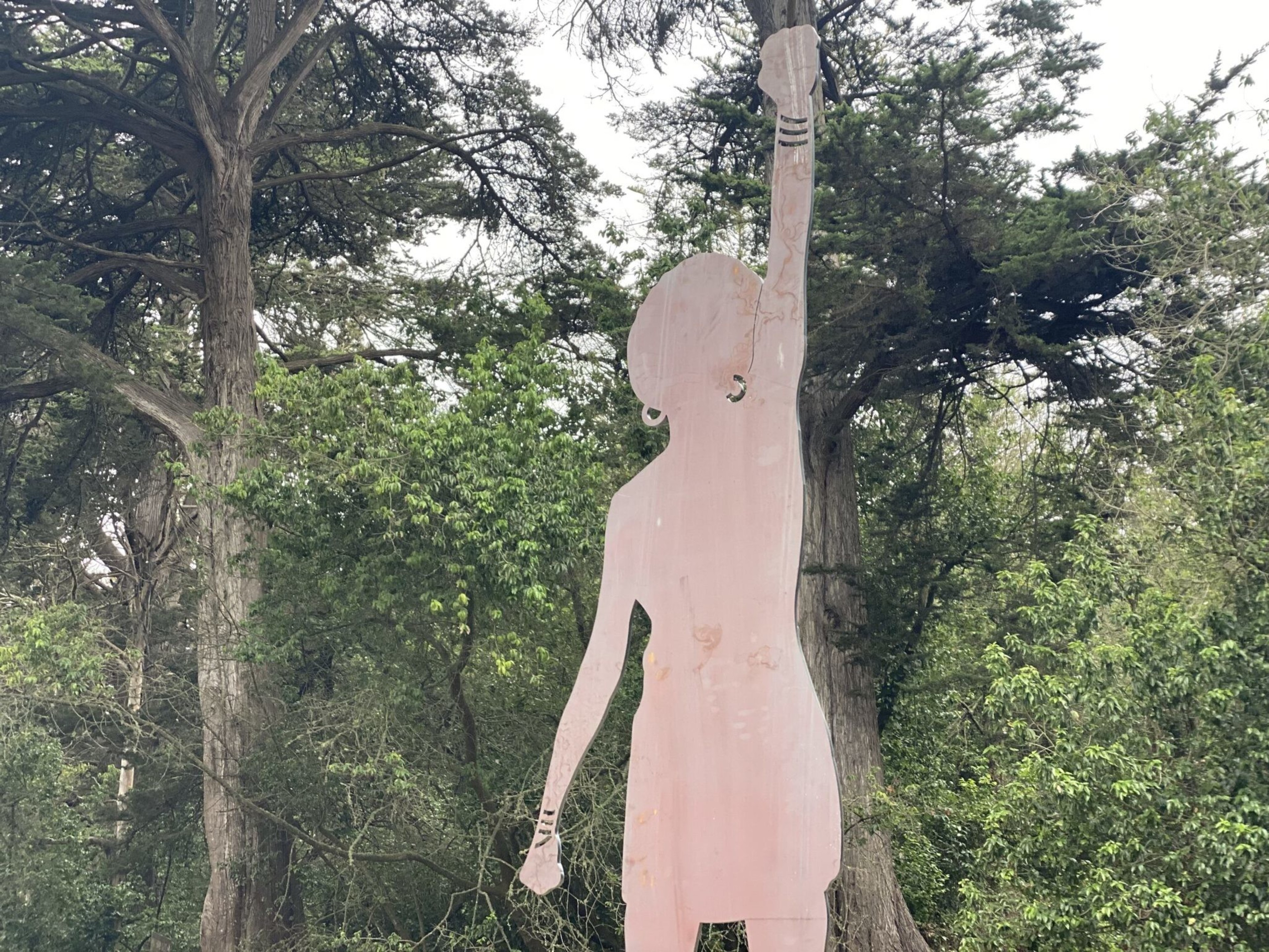 A tall, pink silhouette of a person with one arm raised stands outdoors against a backdrop of green trees and foliage.