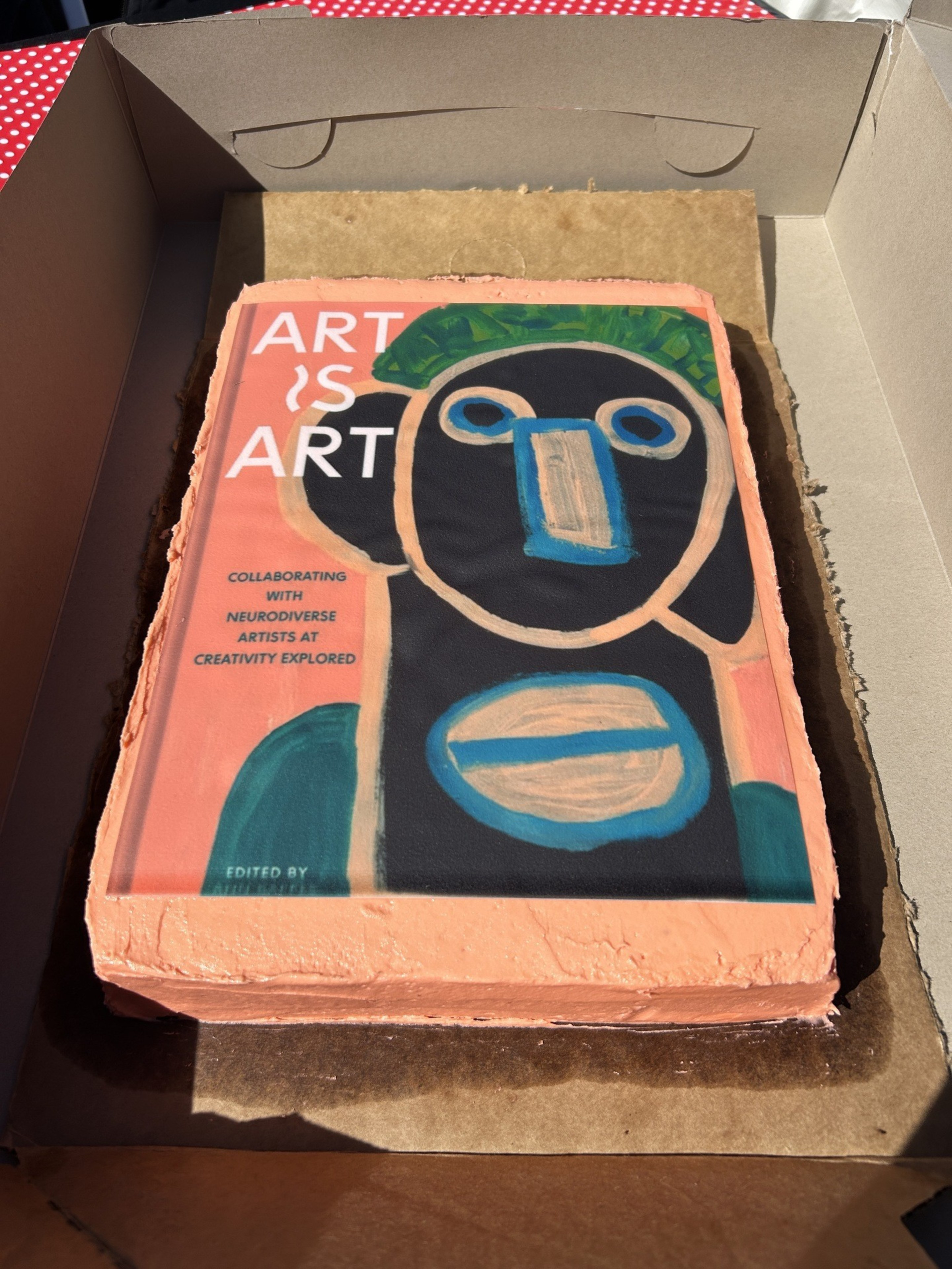 A rectangular cake decorated with orange icing features a colorful abstract face and text reading “ART IS ART” and “Collaborating with neurodiverse artists at Creativity Explored.”
