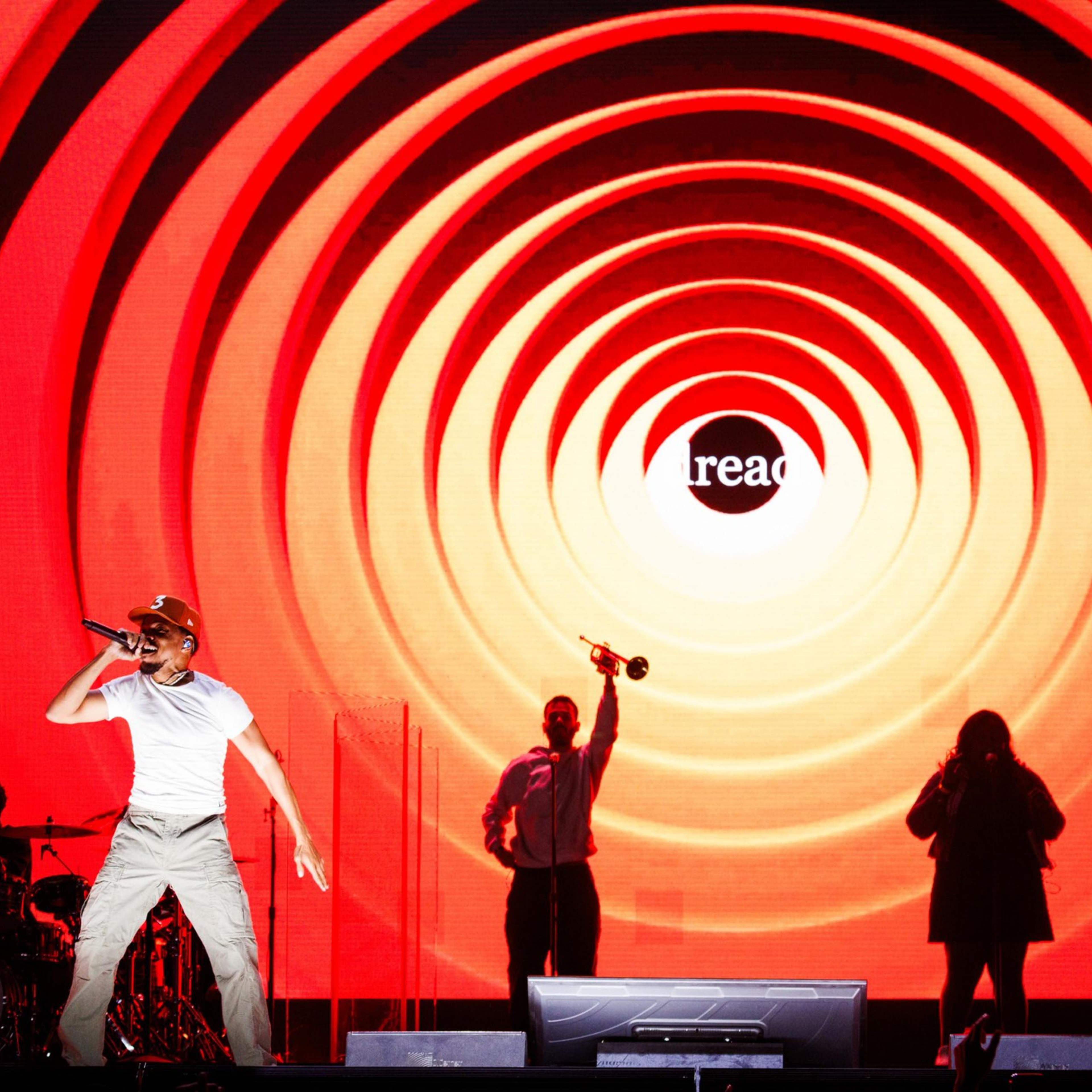 A performer is energetically singing on stage with a red and orange spiral backdrop. Silhouetted band members and a "read" text in the background are also visible.