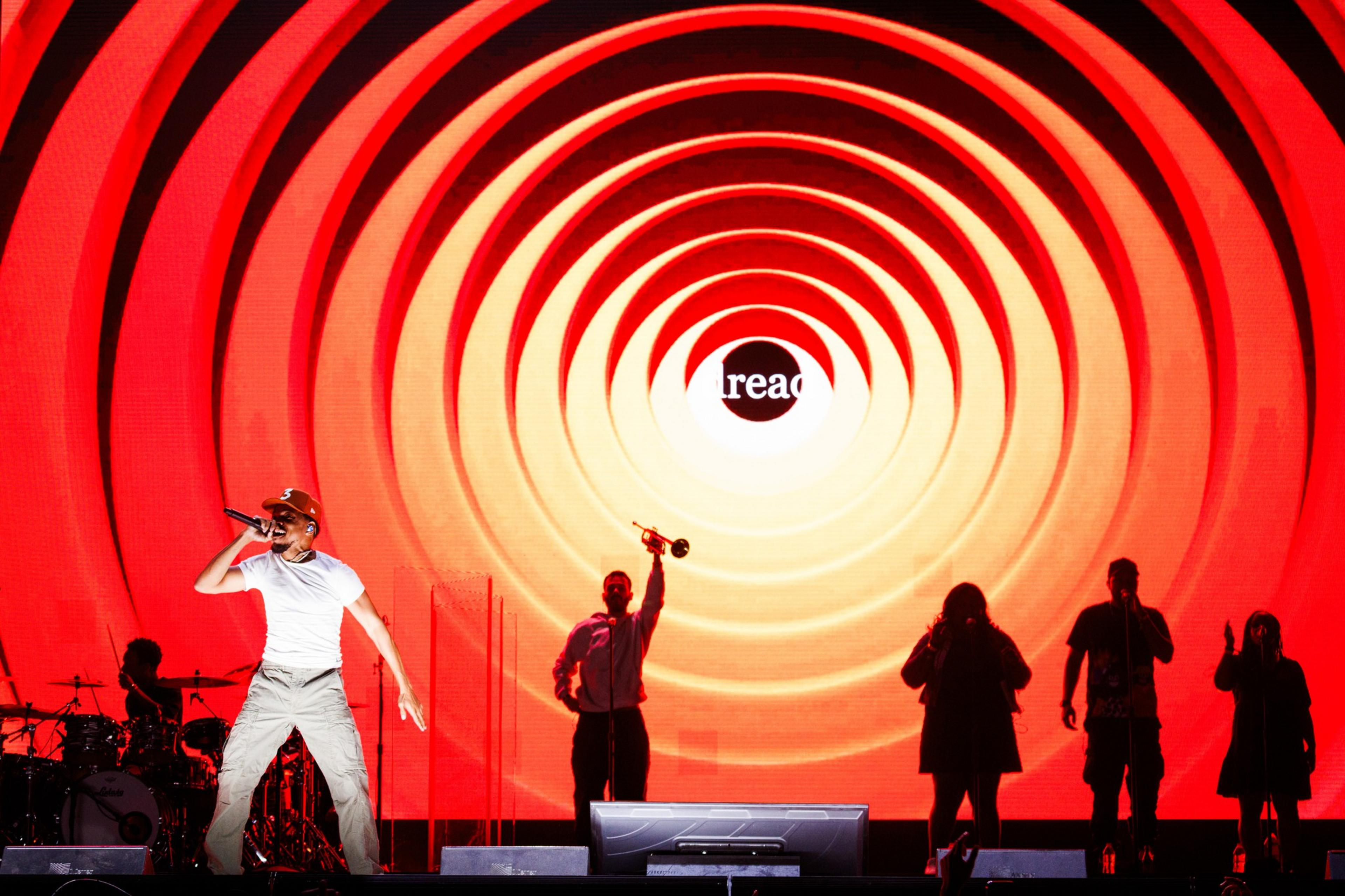 A performer is energetically singing on stage with a red and orange spiral backdrop. Silhouetted band members and a "read" text in the background are also visible.