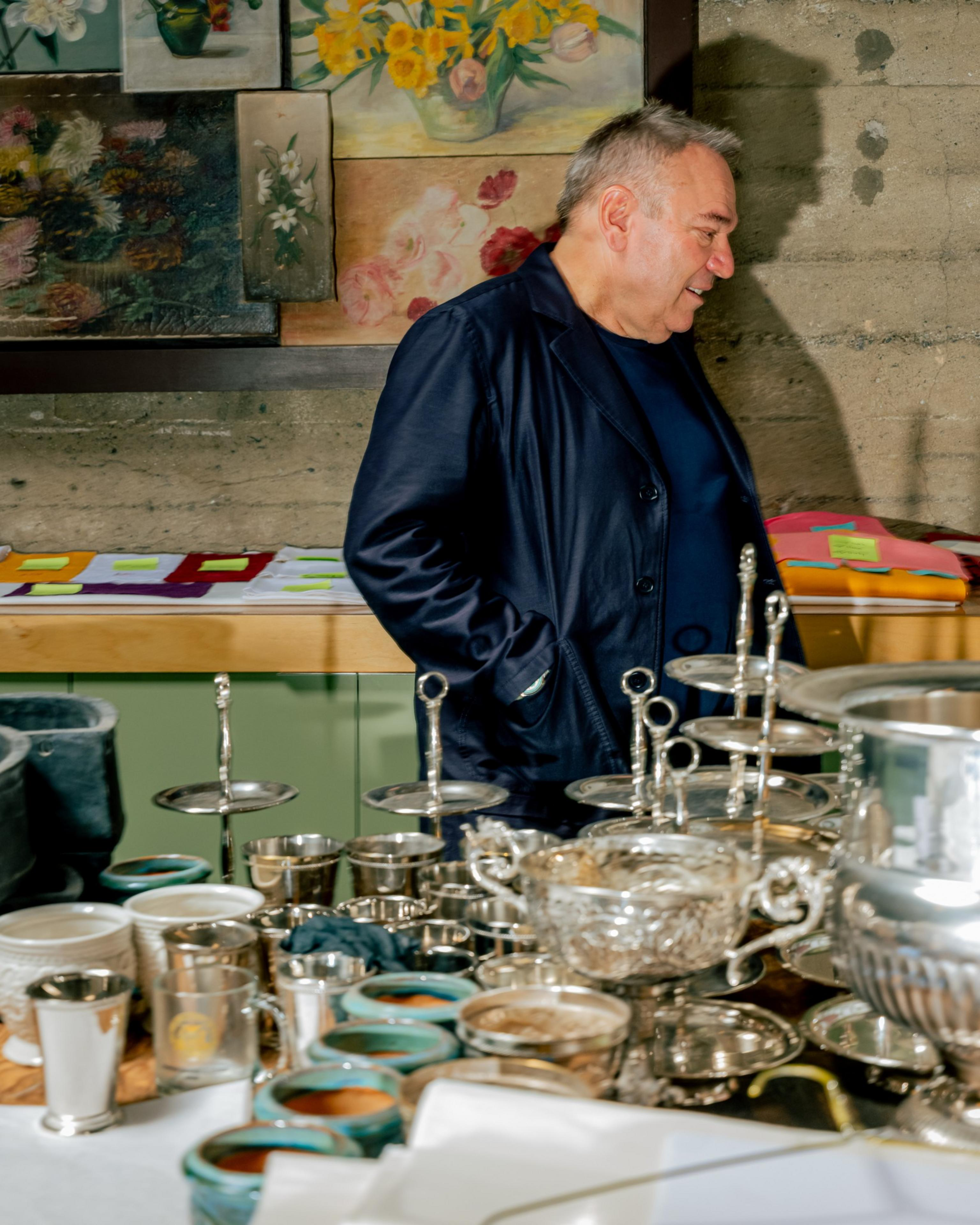 A man in a dark jacket stands next to a table filled with silverware, bowls, candlesticks, and green-rimmed cups, with floral paintings on the wall behind.