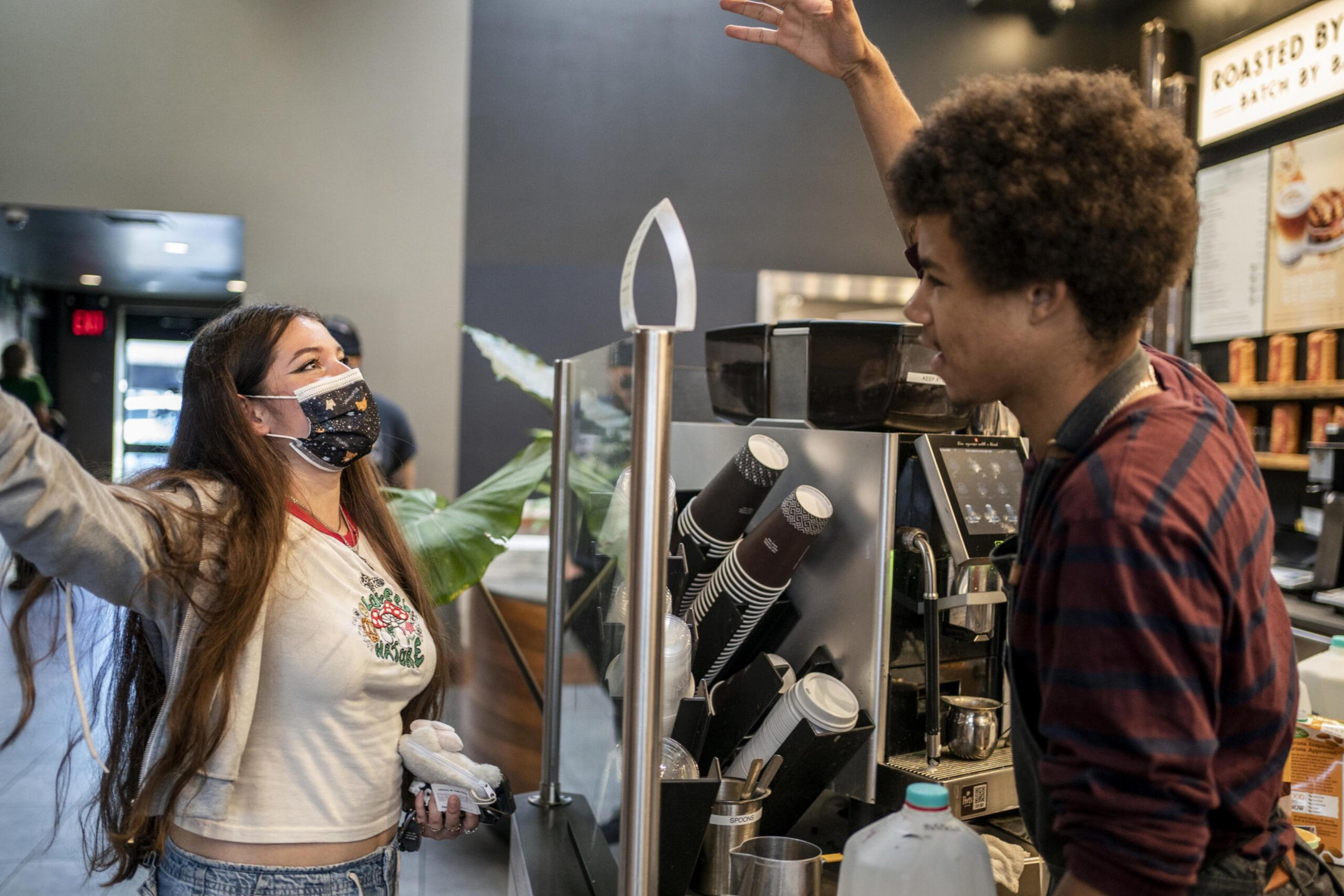 A masked woman with long hair raises her arms joyfully while a smiling barista in a striped shirt chats behind a coffee counter with cups and a milk jug.