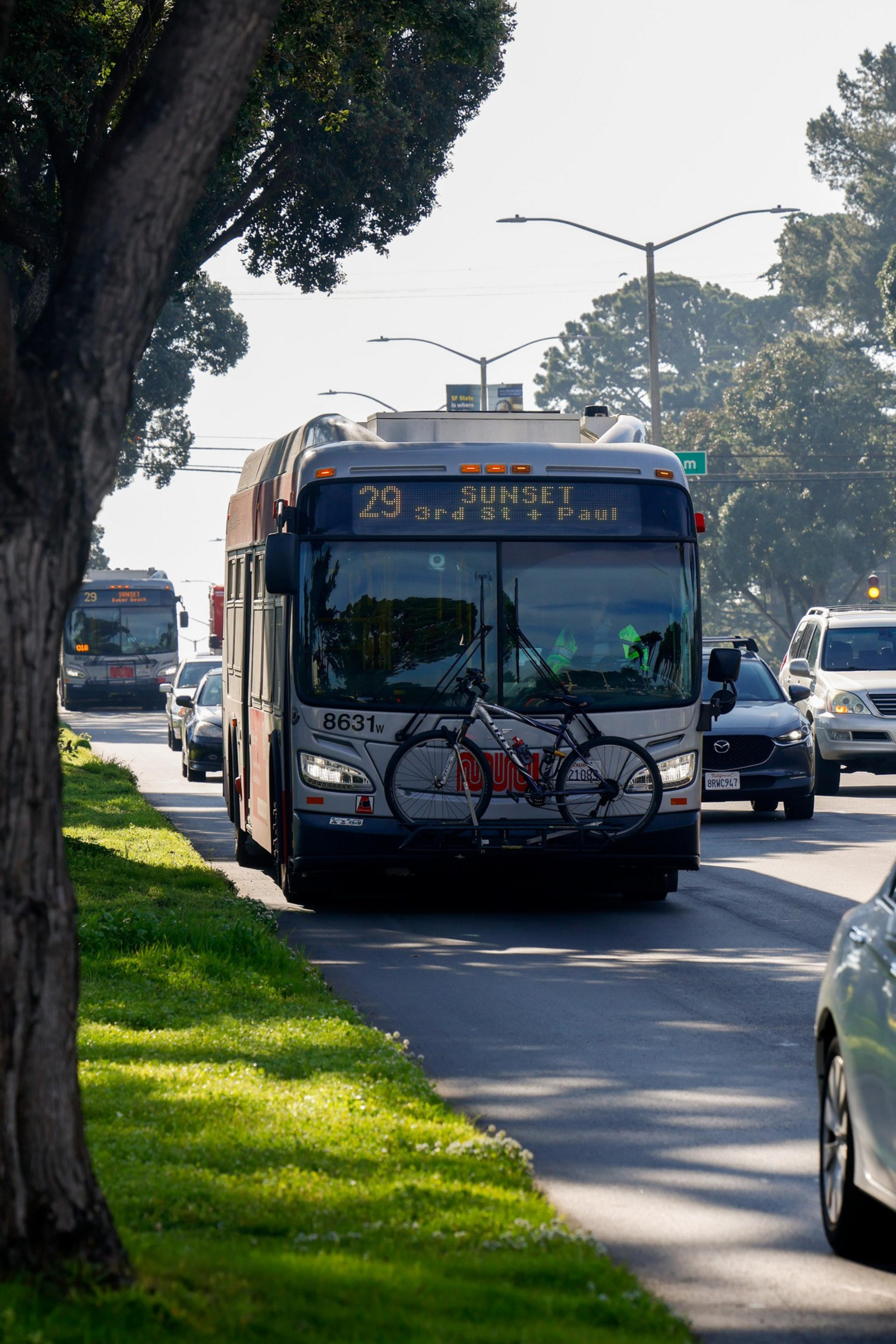A city bus labeled "29 Sunset" with a bicycle mounted on the front drives along a tree-lined street, accompanied by other vehicles.