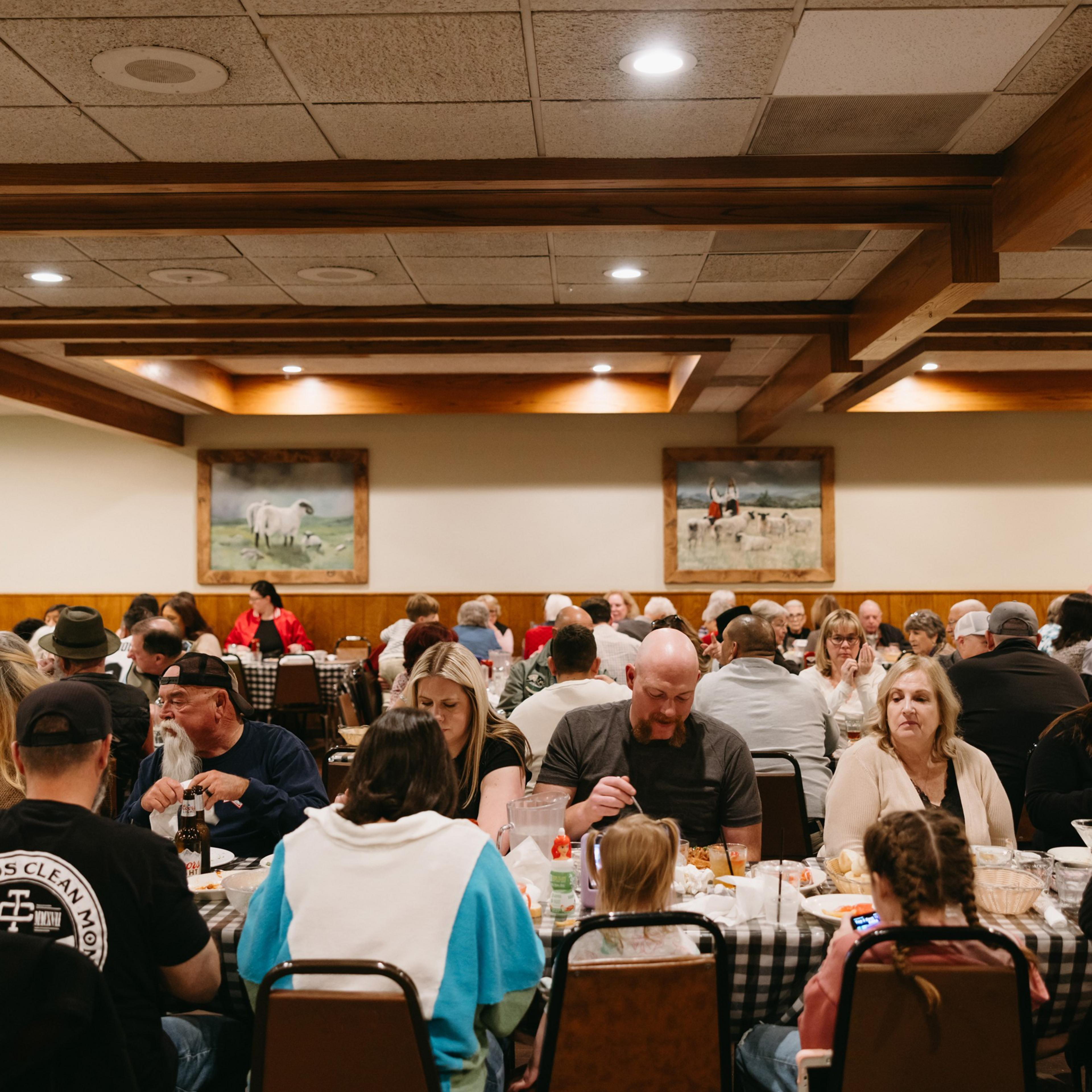 A large group of people is seated at tables in a cozy restaurant, enjoying meals. The room is warmly lit, with framed paintings on the walls.