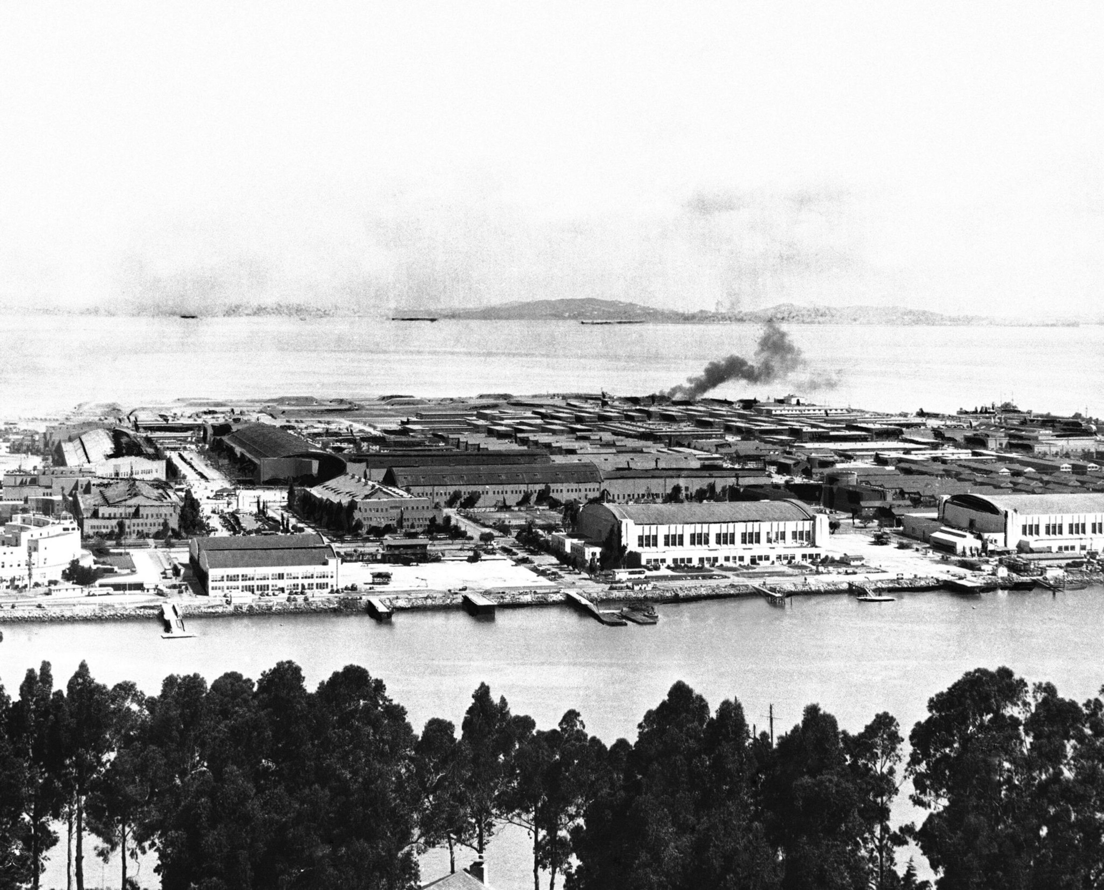 A large industrial complex with multiple warehouses sits by a waterfront, with trees in the foreground and a coastline in the distance under a smoky sky.