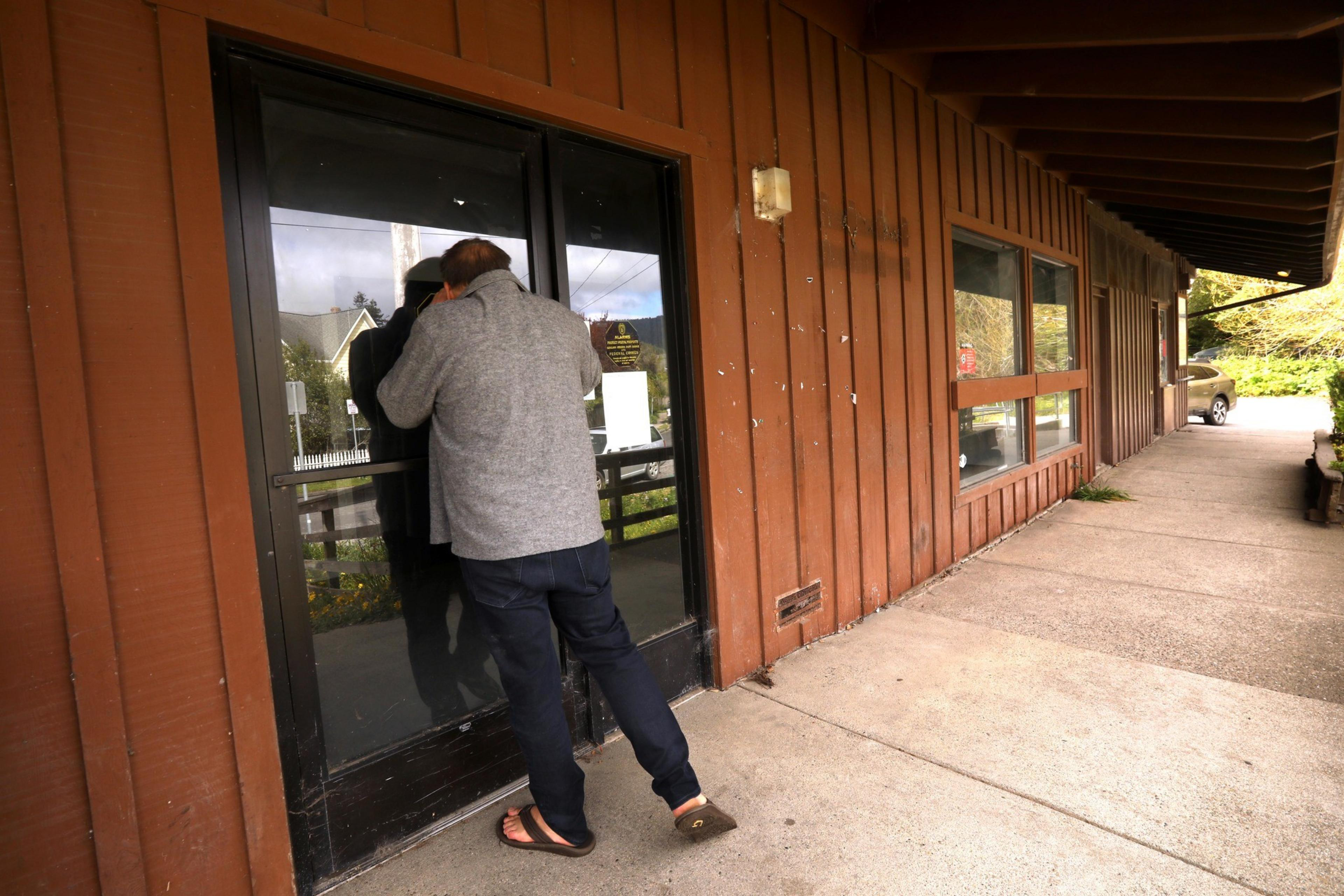 A person wearing a gray jacket and jeans is peering through the glass door of a brown wooden building. A car is partially visible in the distance.