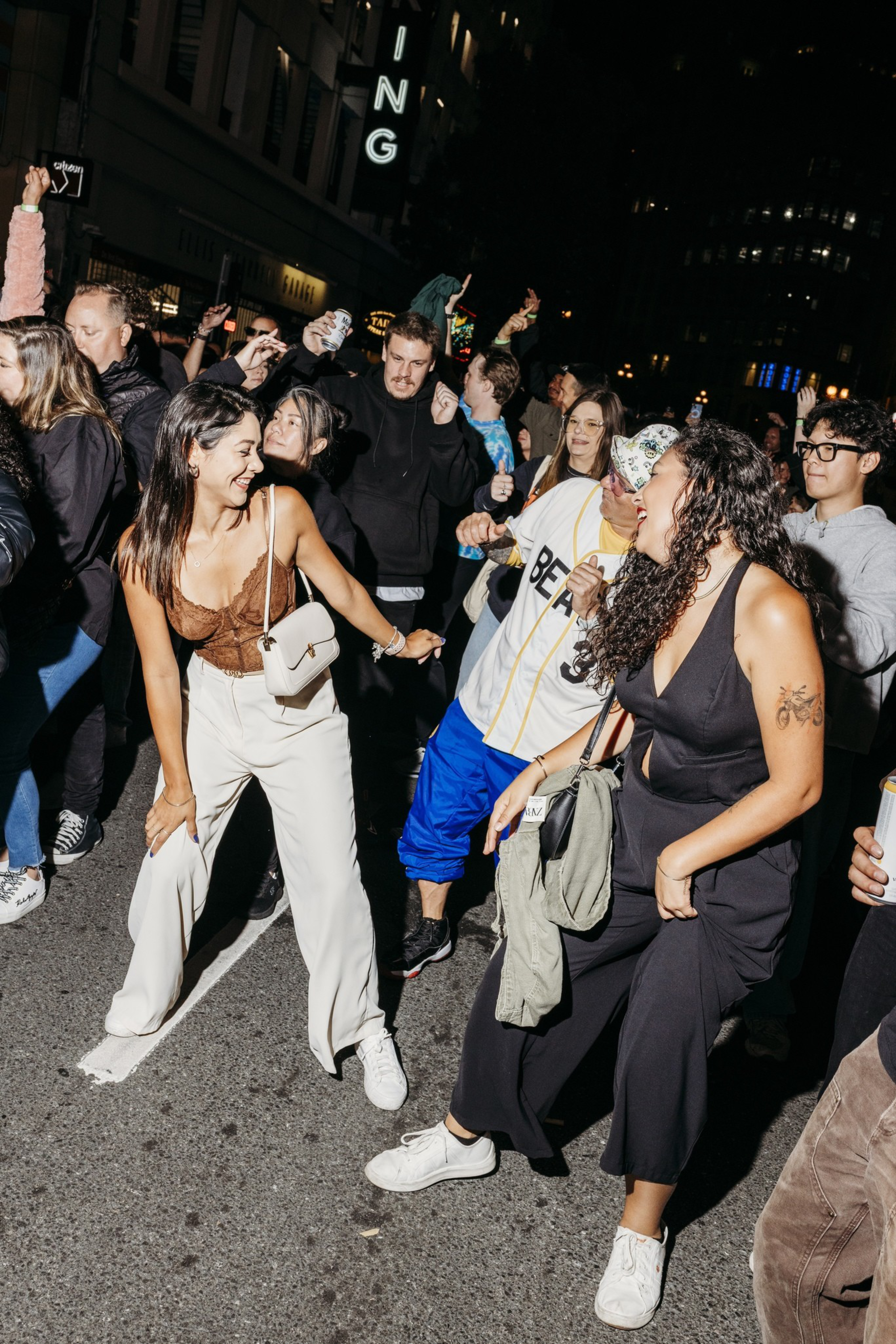 People are dancing and enjoying a lively street party at night, with smiles, casual outfits, and hands raised in celebration.
