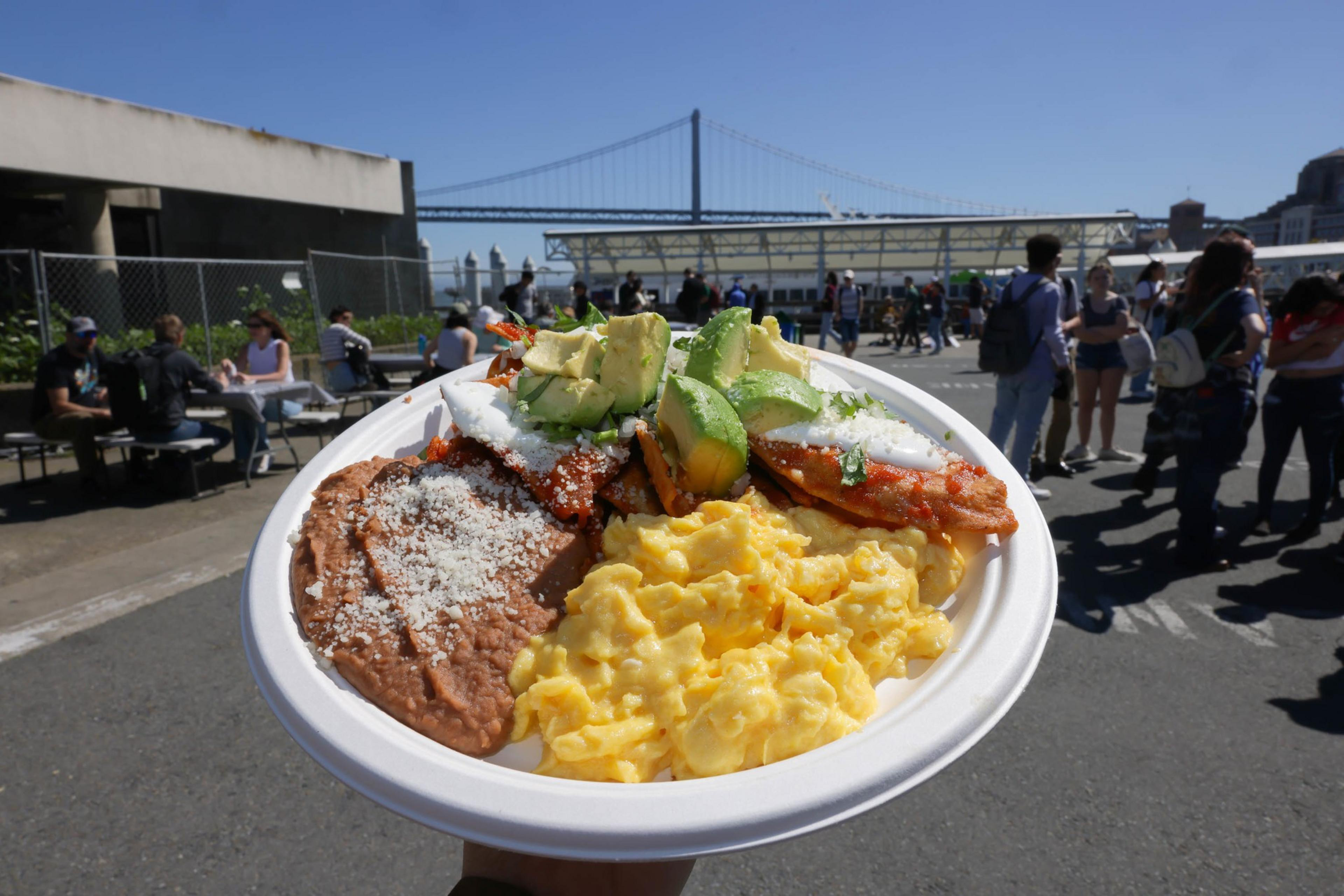 A plate of food with eggs, beans, avocado, and bacon, with a bridge and people in the background.