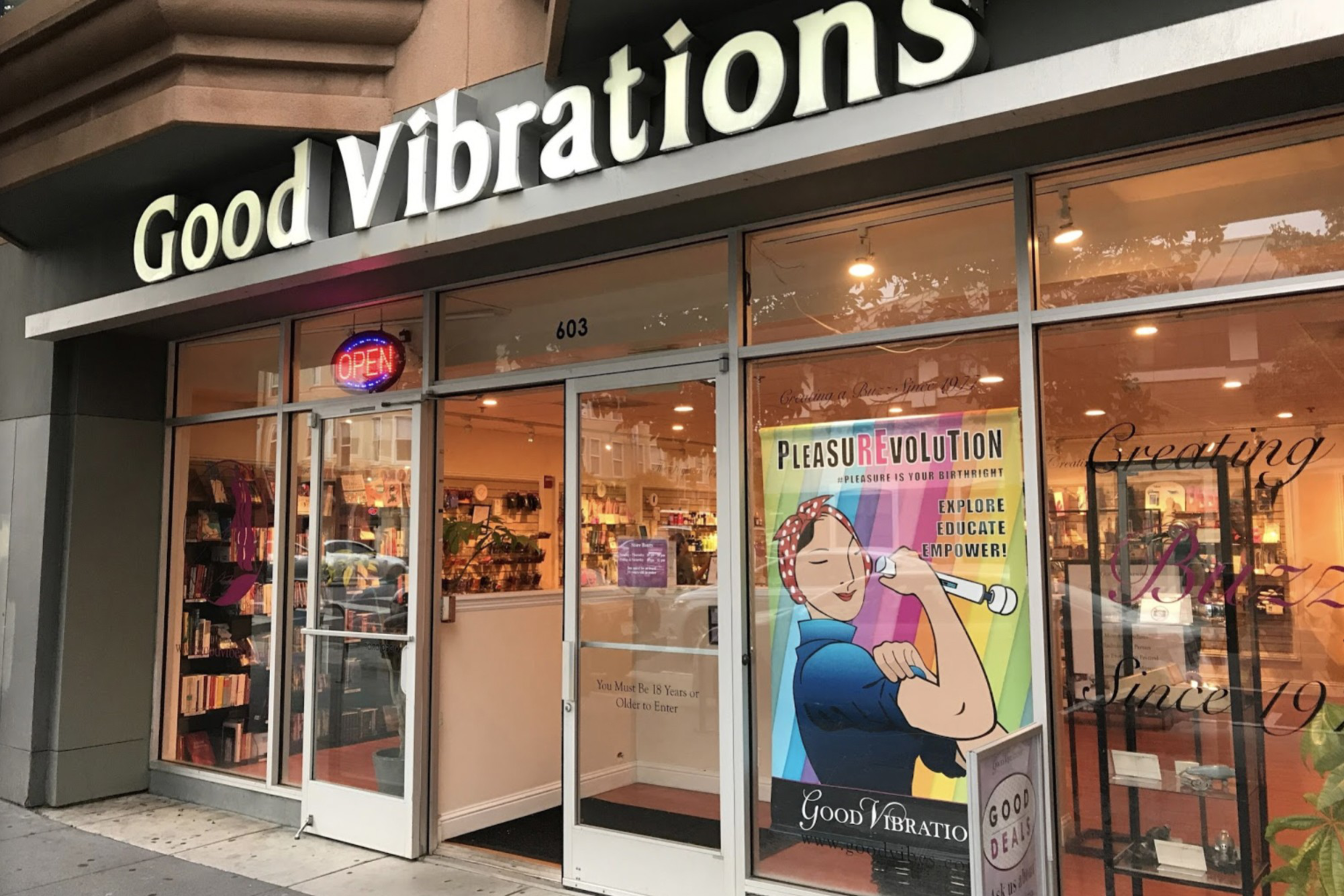 The image shows the storefront of &quot;Good Vibrations,&quot; with a neon open sign and a colorful poster promoting a &quot;Pleasure Revolution&quot; inside the glass door.