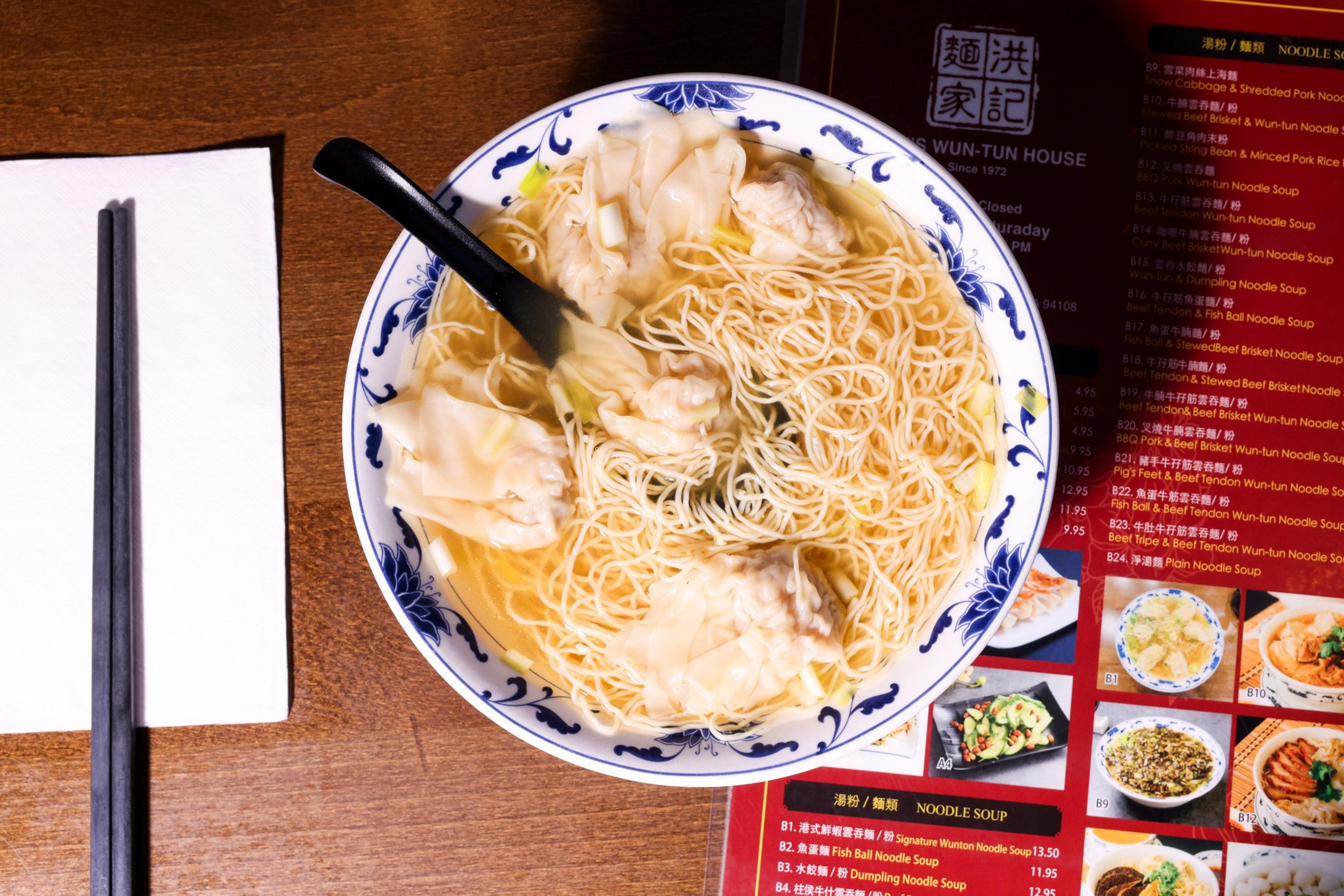 A bowl of noodle soup with dumplings sits on a wooden table, accompanied by chopsticks and a menu nearby. A napkin and a black spoon are also visible.
