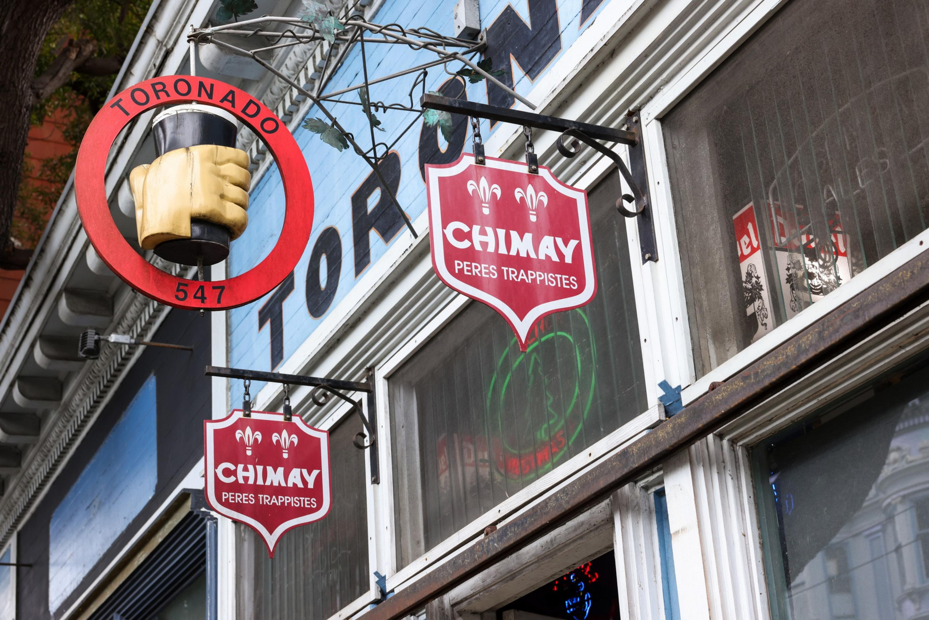 A storefront with a red circular sign featuring a hand holding a mug, labeled &quot;TORONADO 547,&quot; and a red shield sign reading &quot;CHIMAY PERES TRAPPISTES.&quot;