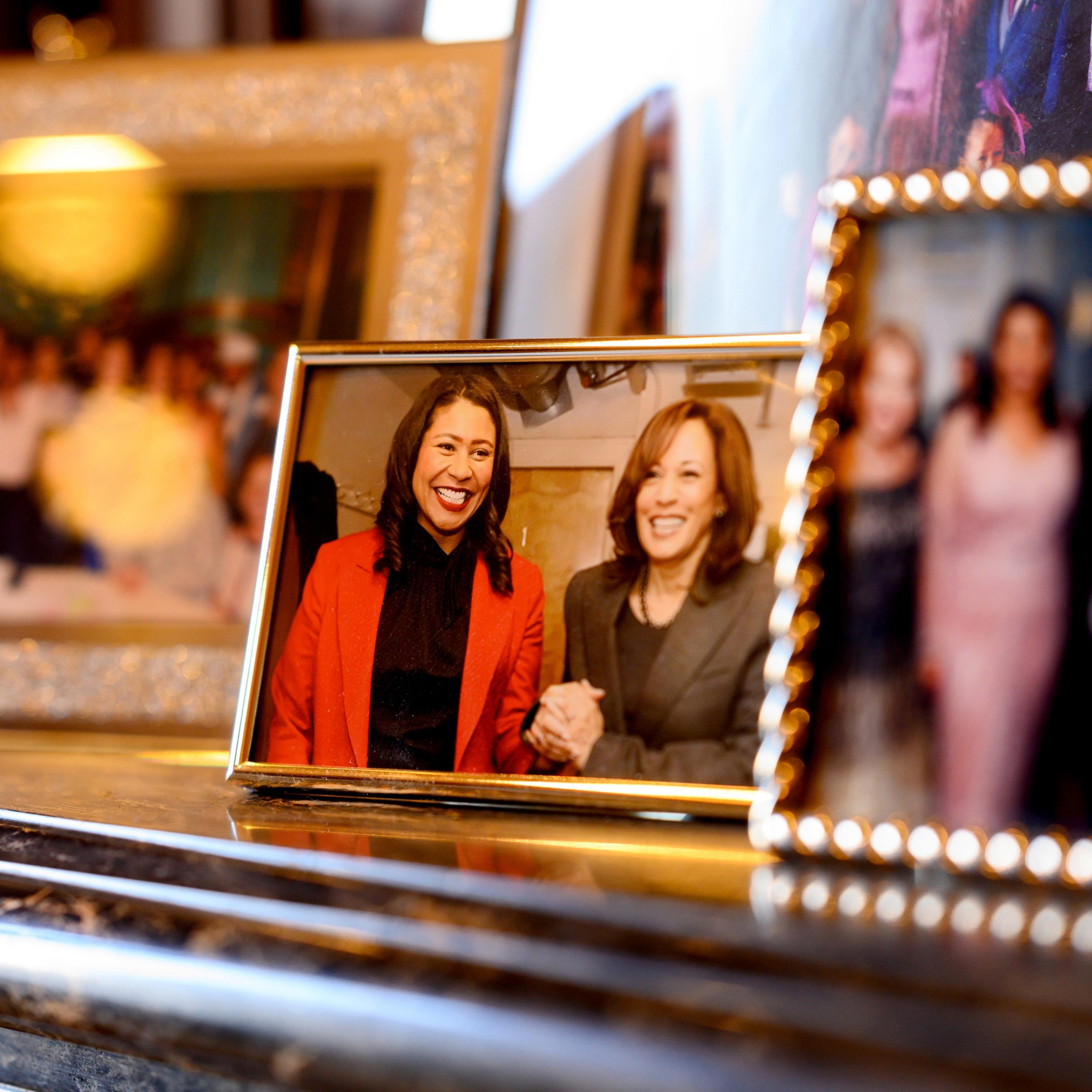 The image shows several framed photographs with the central one featuring two smiling women holding hands, dressed in professional attire. The setting appears to be indoors.