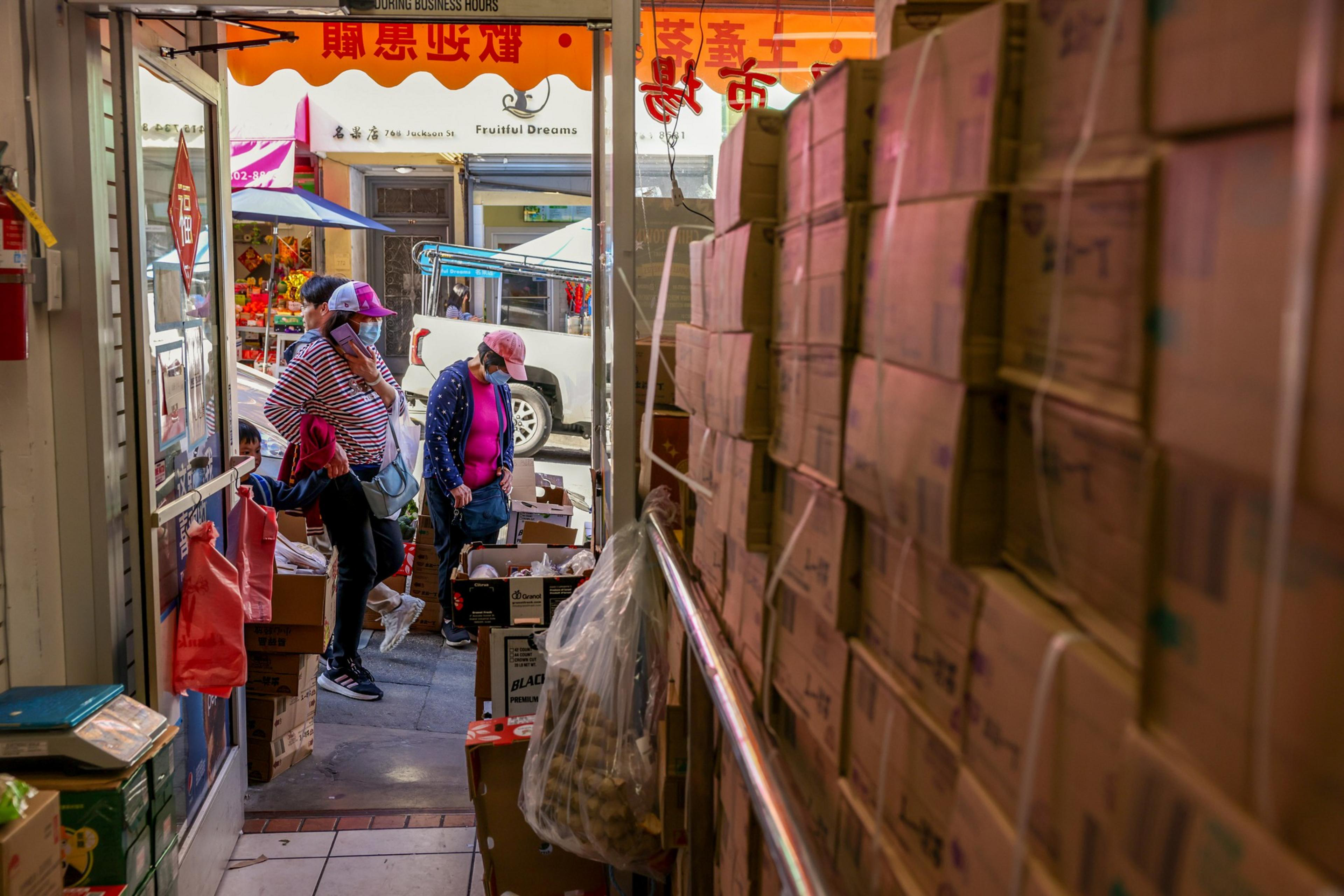 A bustling store entrance shows people in hats examining boxes, with tall stacks on the right and colorful signs outside, suggesting a busy market.