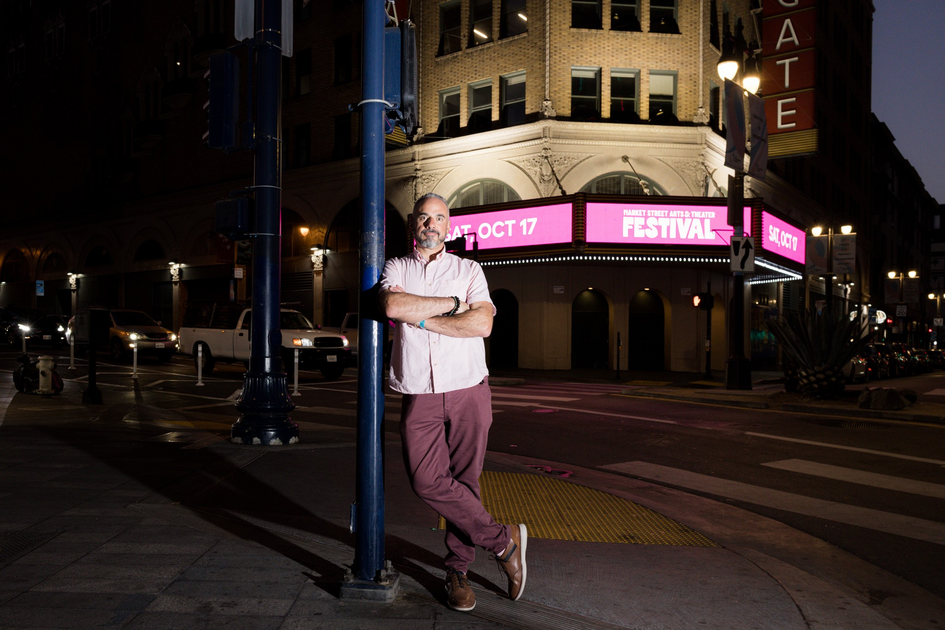 A man with a beard leans against a street pole at night near a theater with a bright marquee advertising a festival on