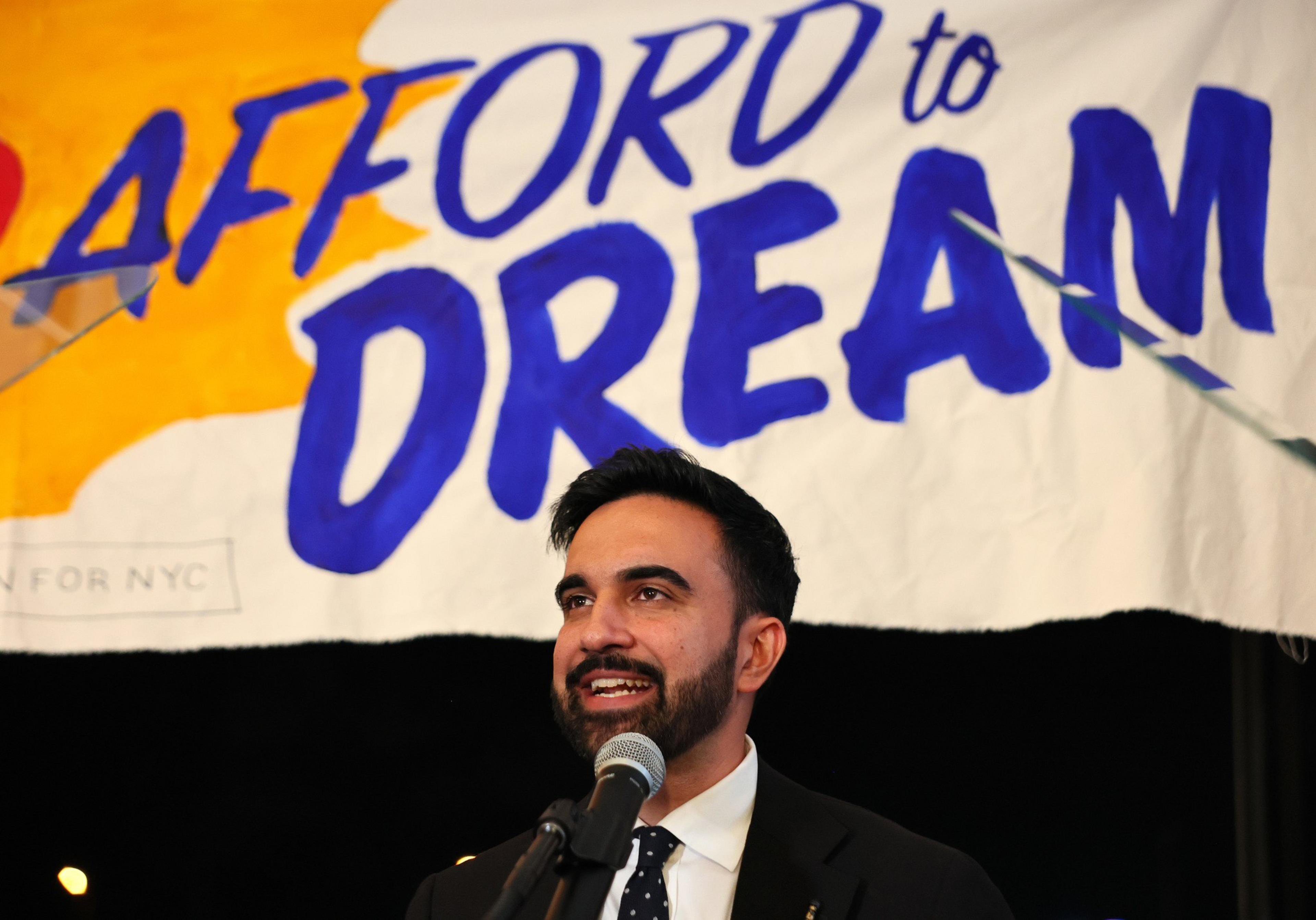 A man stands in front of a microphone, smiling, with a banner behind him reading "AFFORD to DREAM" in large, colorful letters.