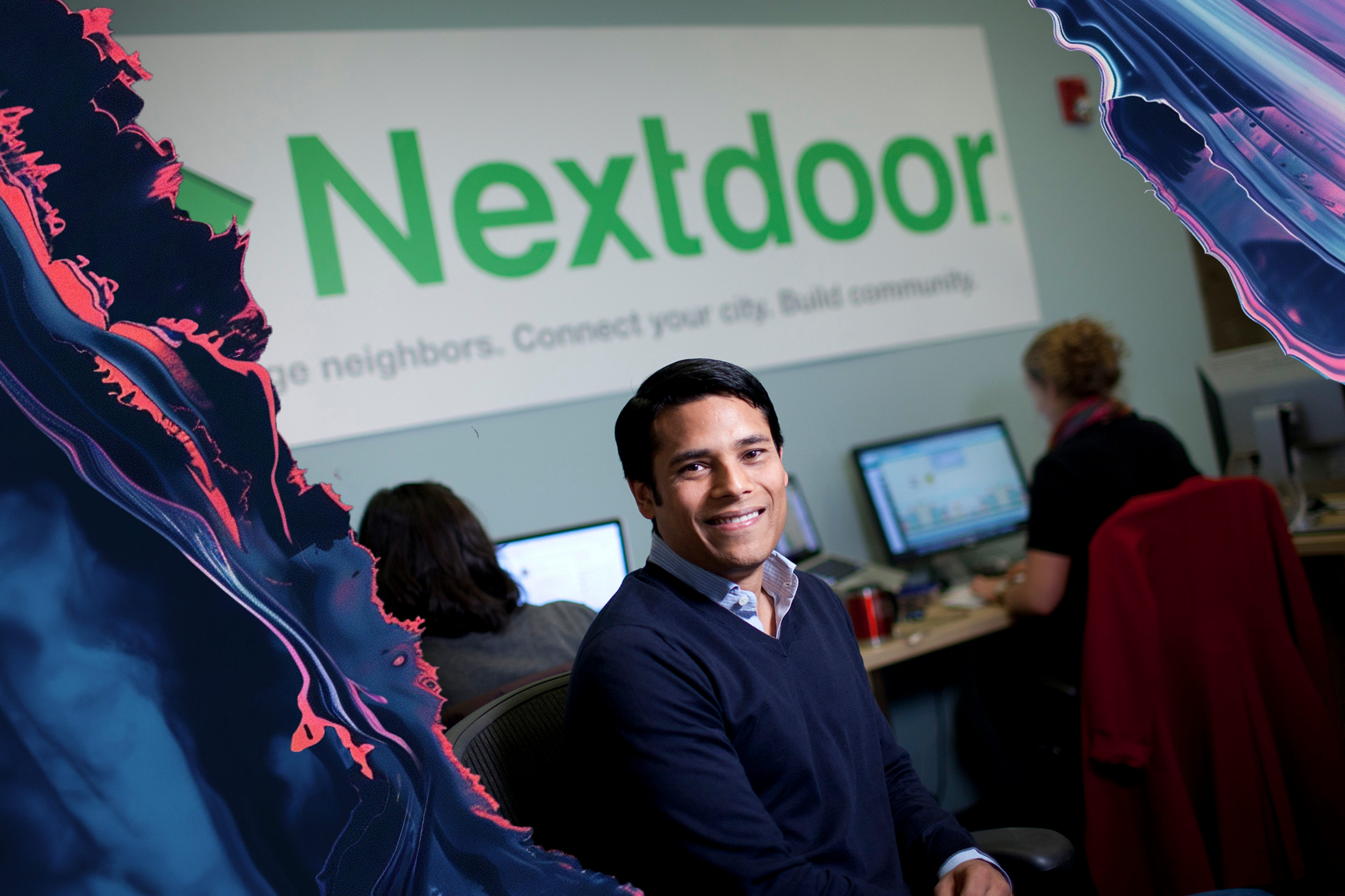 A man is smiling and sitting in an office with two others working on computers. The word "Nextdoor" is prominently displayed on a wall behind them.