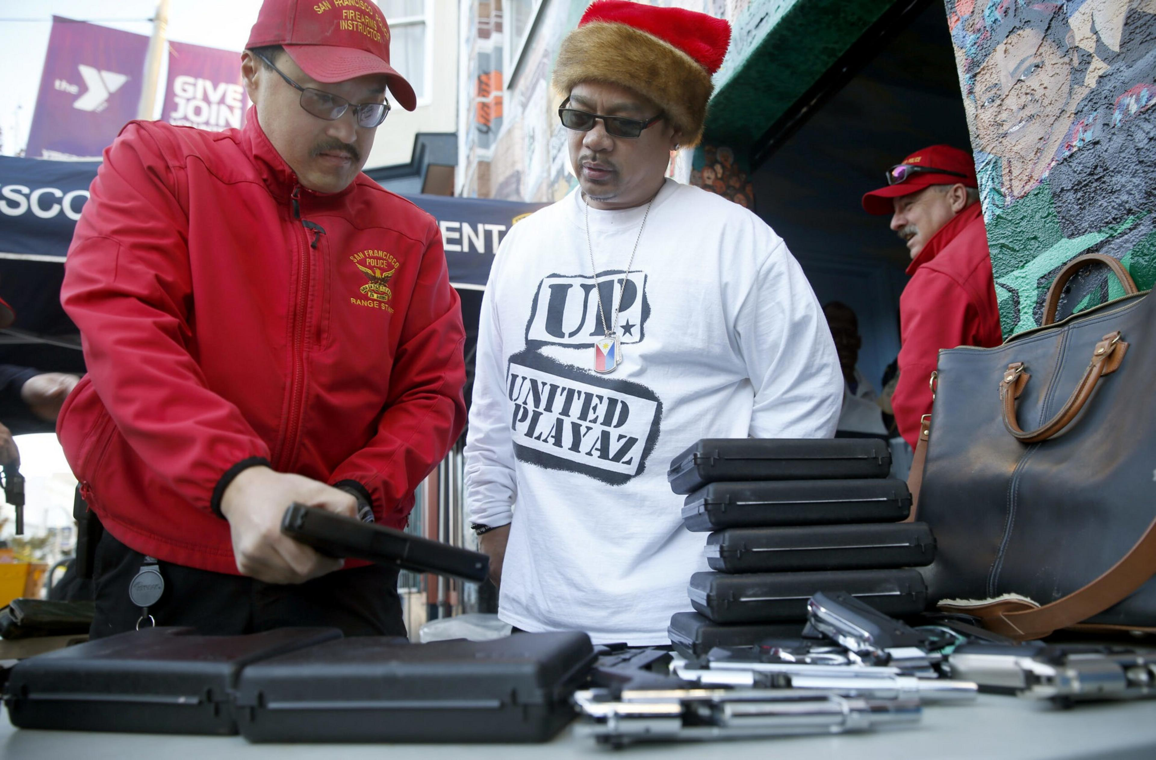 A man in a red jacket and cap inspects a handgun on a table filled with gun cases and pistols while another man in a white shirt and red hat watches.