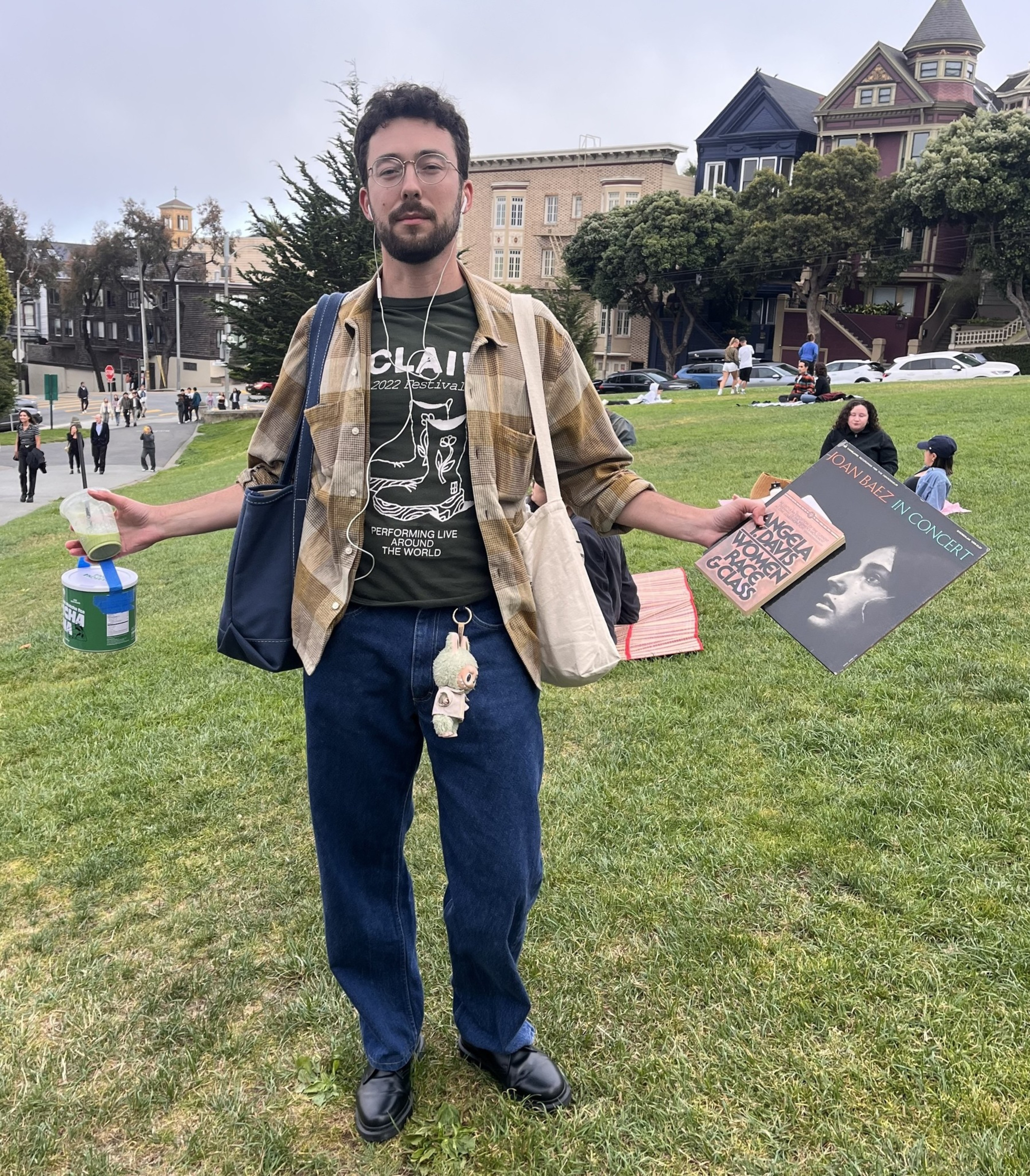 A man stands on grass holding a green drink and two vinyl records, wearing glasses, a plaid shirt, dark pants, black shoes, and carrying two tote bags.
