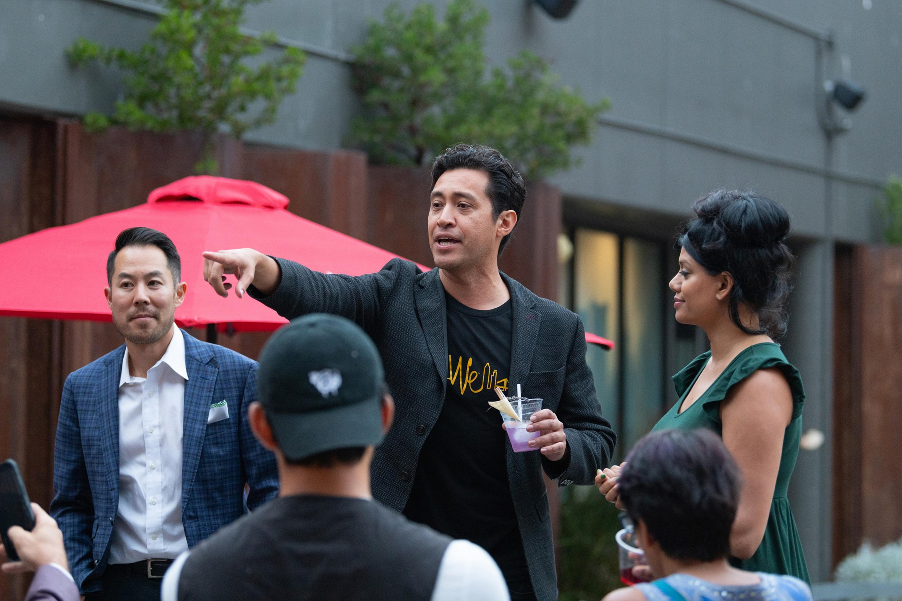 A man in a black jacket is speaking and pointing while holding a drink, surrounded by two men and a woman outdoors near a red umbrella.