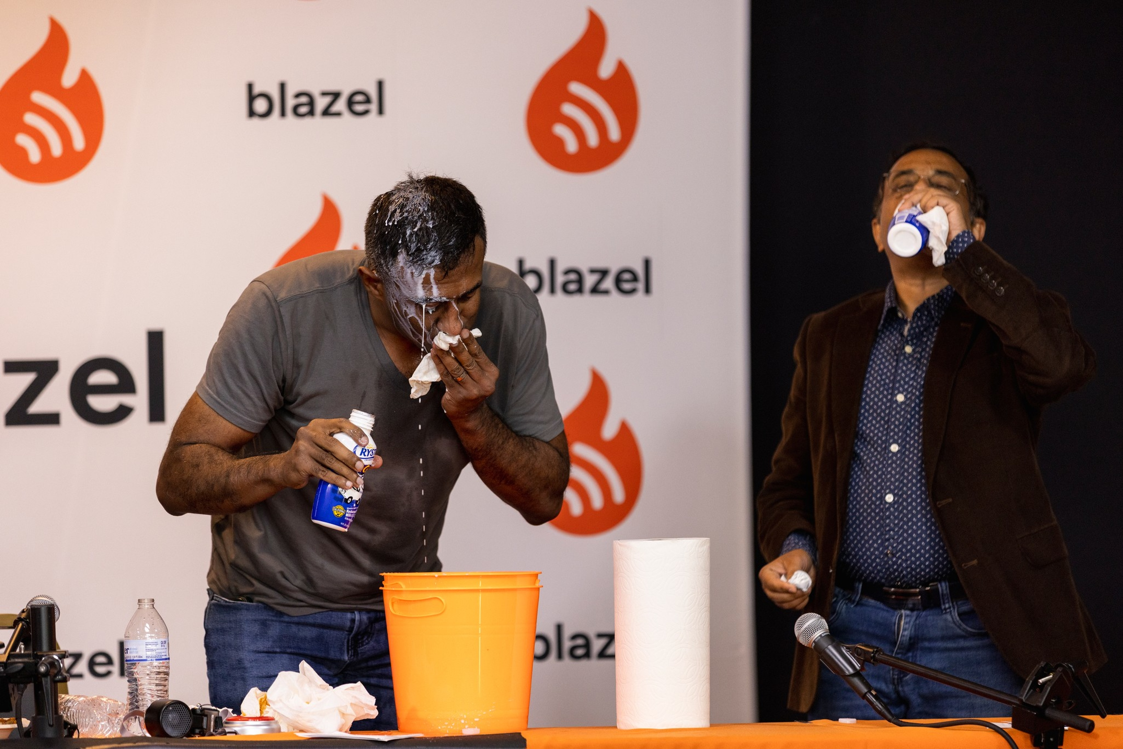 Two men drink from small bottles while standing behind a table with an orange bucket, paper towels, and a microphone, against a backdrop with “blazel” logos.