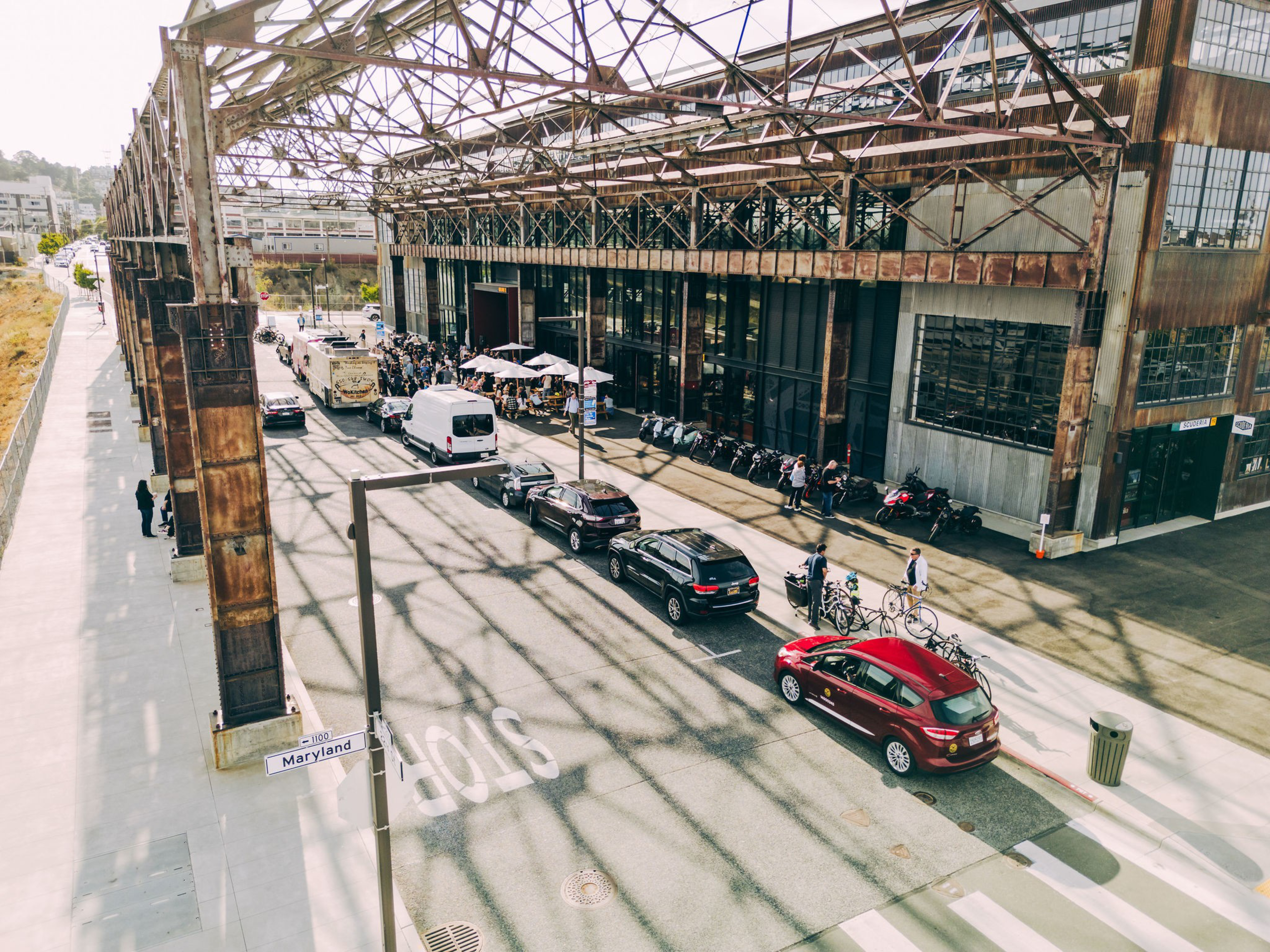 The image shows a busy street next to an industrial-style building with cars, a van, and people walking. Bicycles and motorcycles are lined up outside.