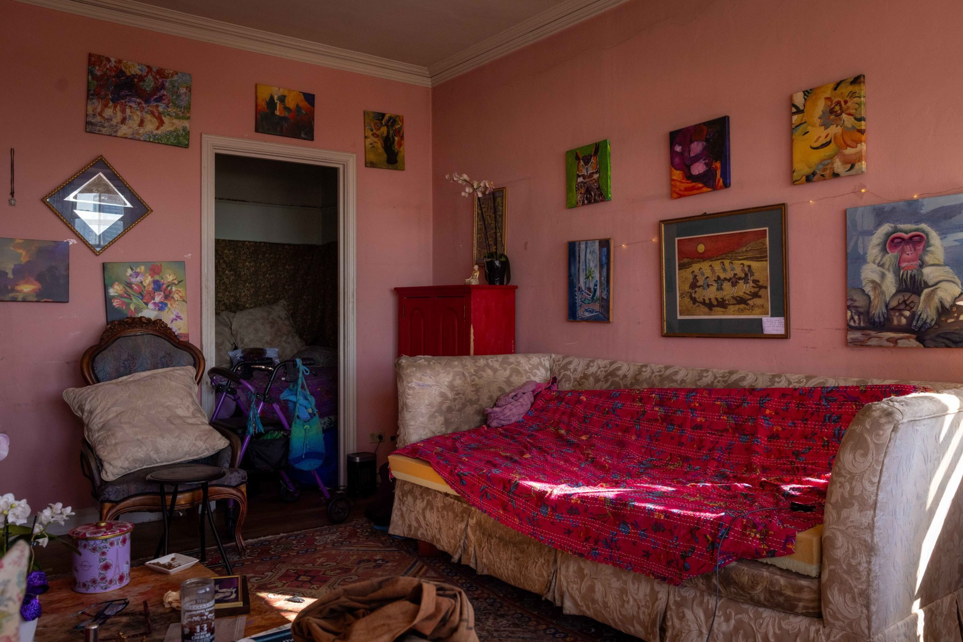A cozy room with pink walls features a beige sofa draped with a red blanket, various colorful paintings, a cushioned chair, and a red cabinet near a doorway.