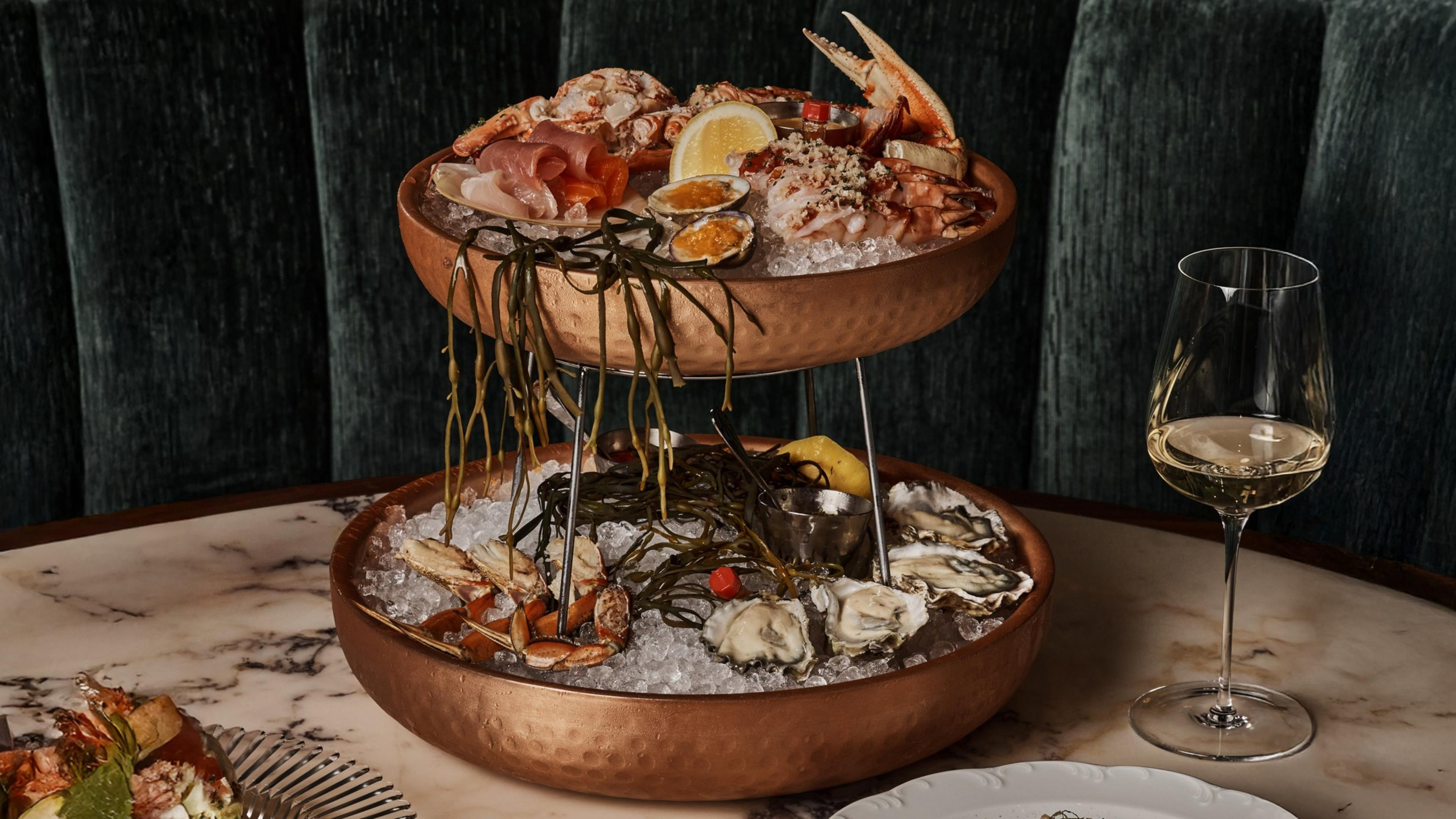 A two-tiered copper seafood platter filled with ice, oysters, crab, shrimp, lemon wedges, seaweed, and a glass of white wine on the side.