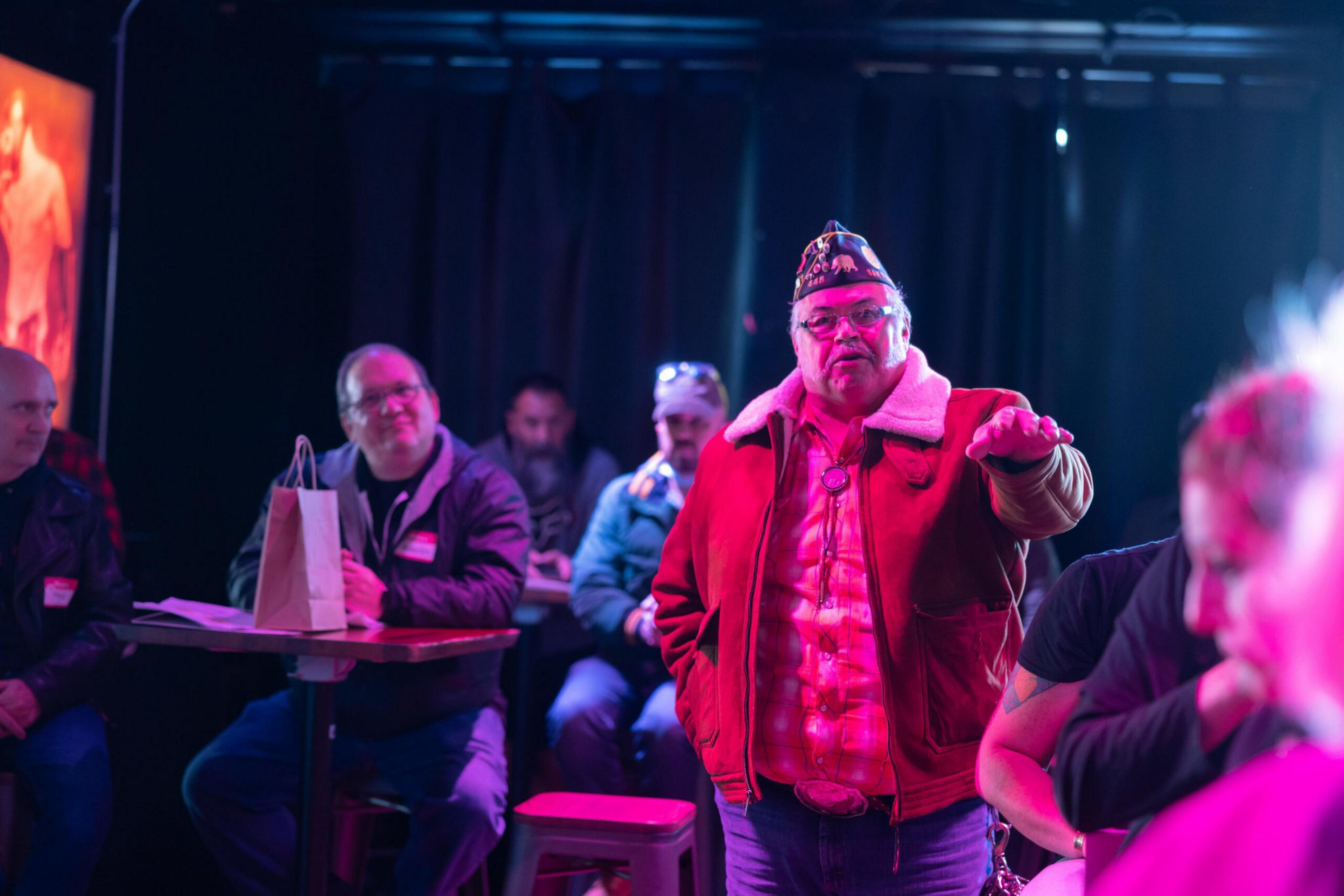 A man wearing a red jacket and a decorated hat speaks to a small group seated in a dimly lit room with purple and pink lighting.