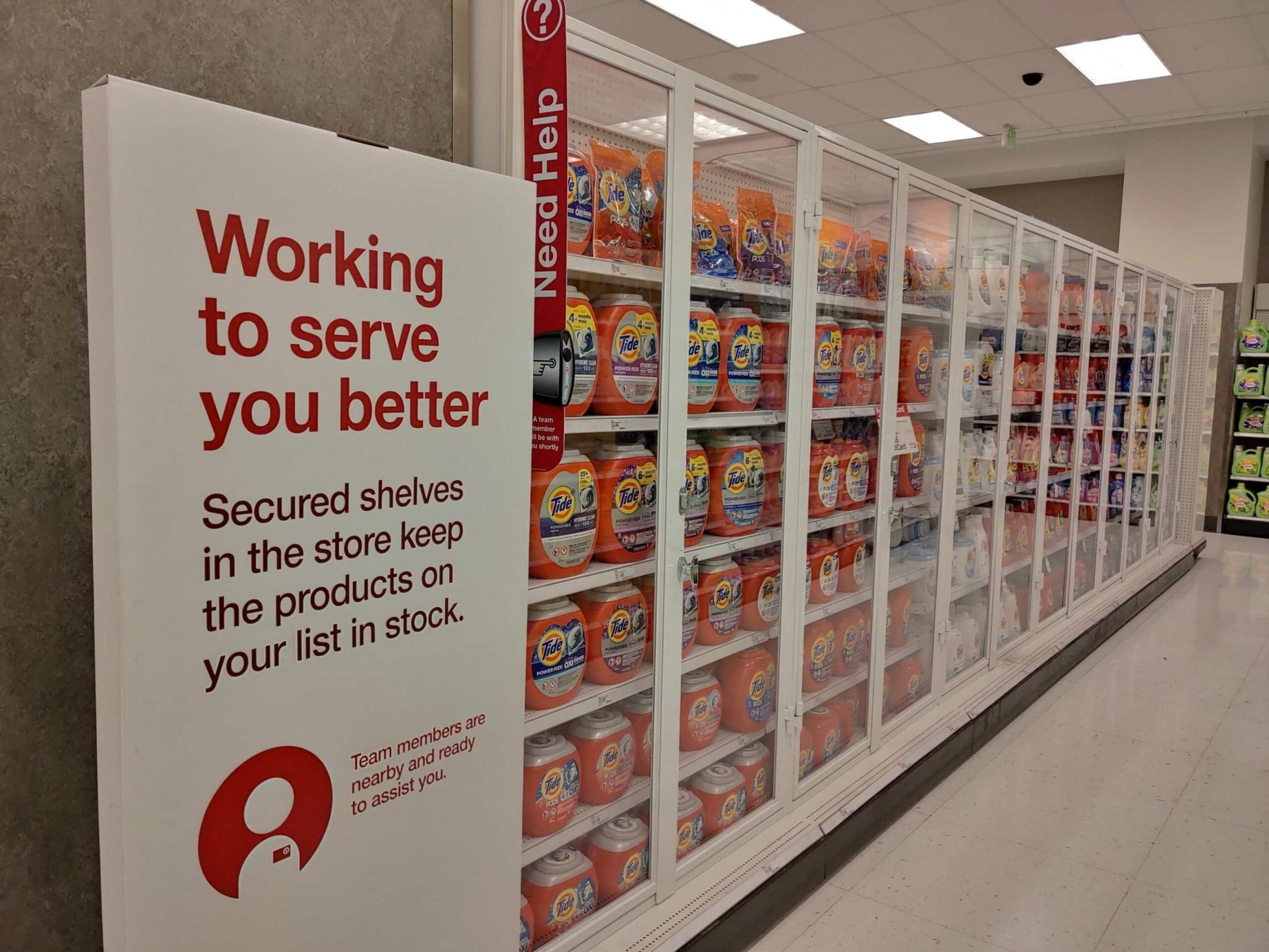 Secured glass shelves in a store display multiple detergent bottles behind locked doors, with a sign explaining the shelves keep products in stock for customers.