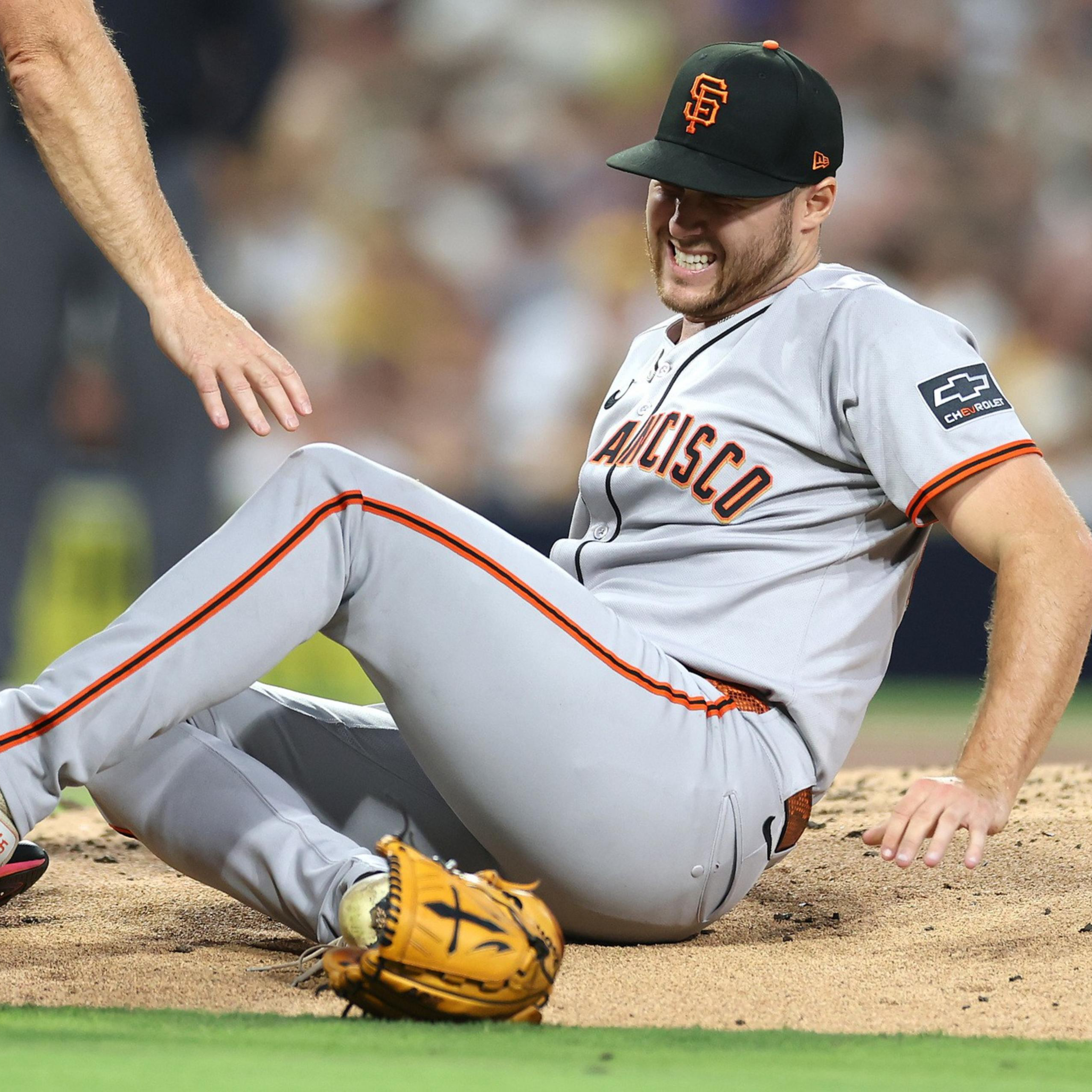 A San Francisco Giants baseball player is sitting on the ground, grimacing in pain, while an umpire reaches toward him.