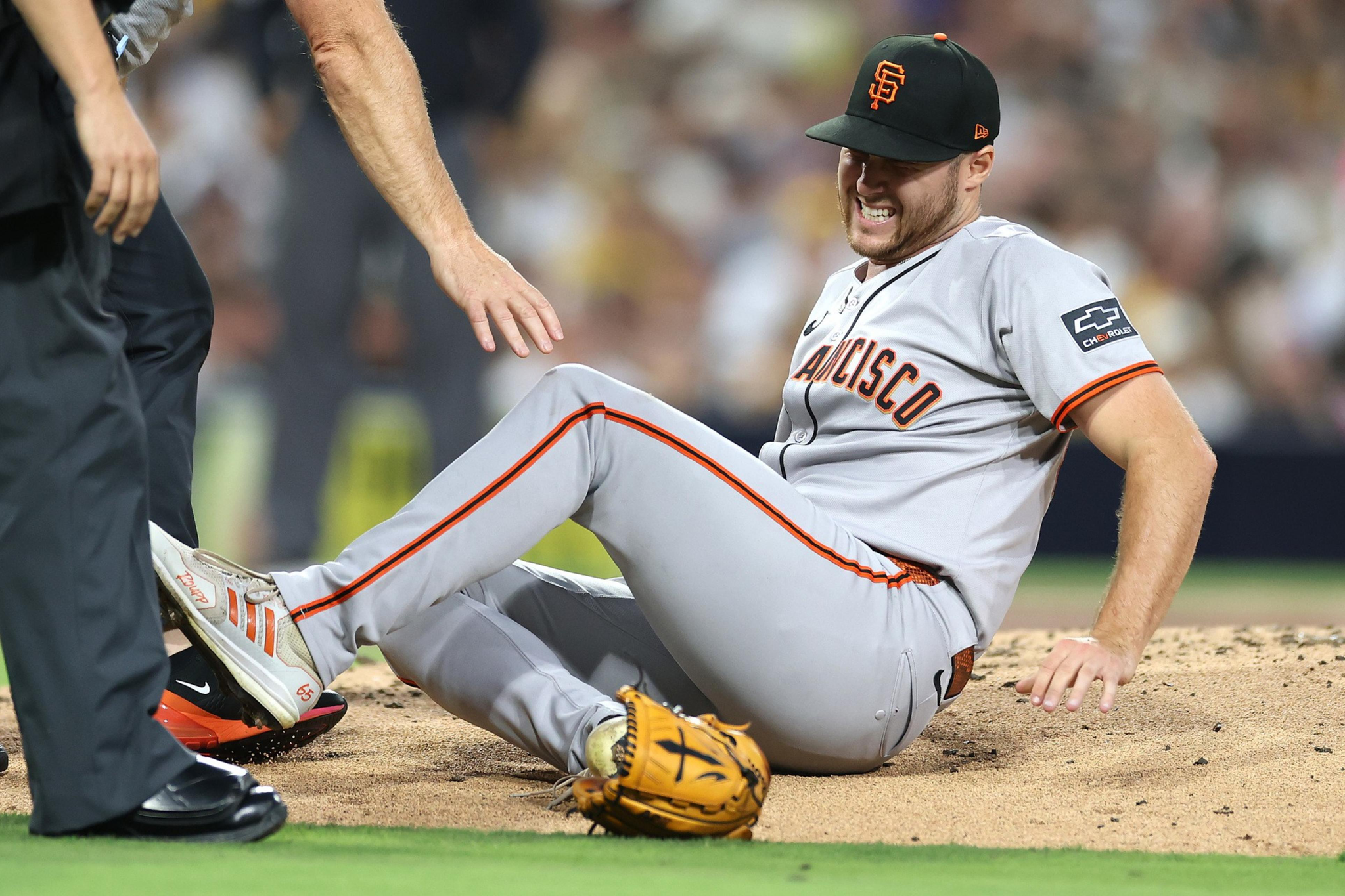 A San Francisco Giants baseball player is sitting on the ground, grimacing in pain, while an umpire reaches toward him.