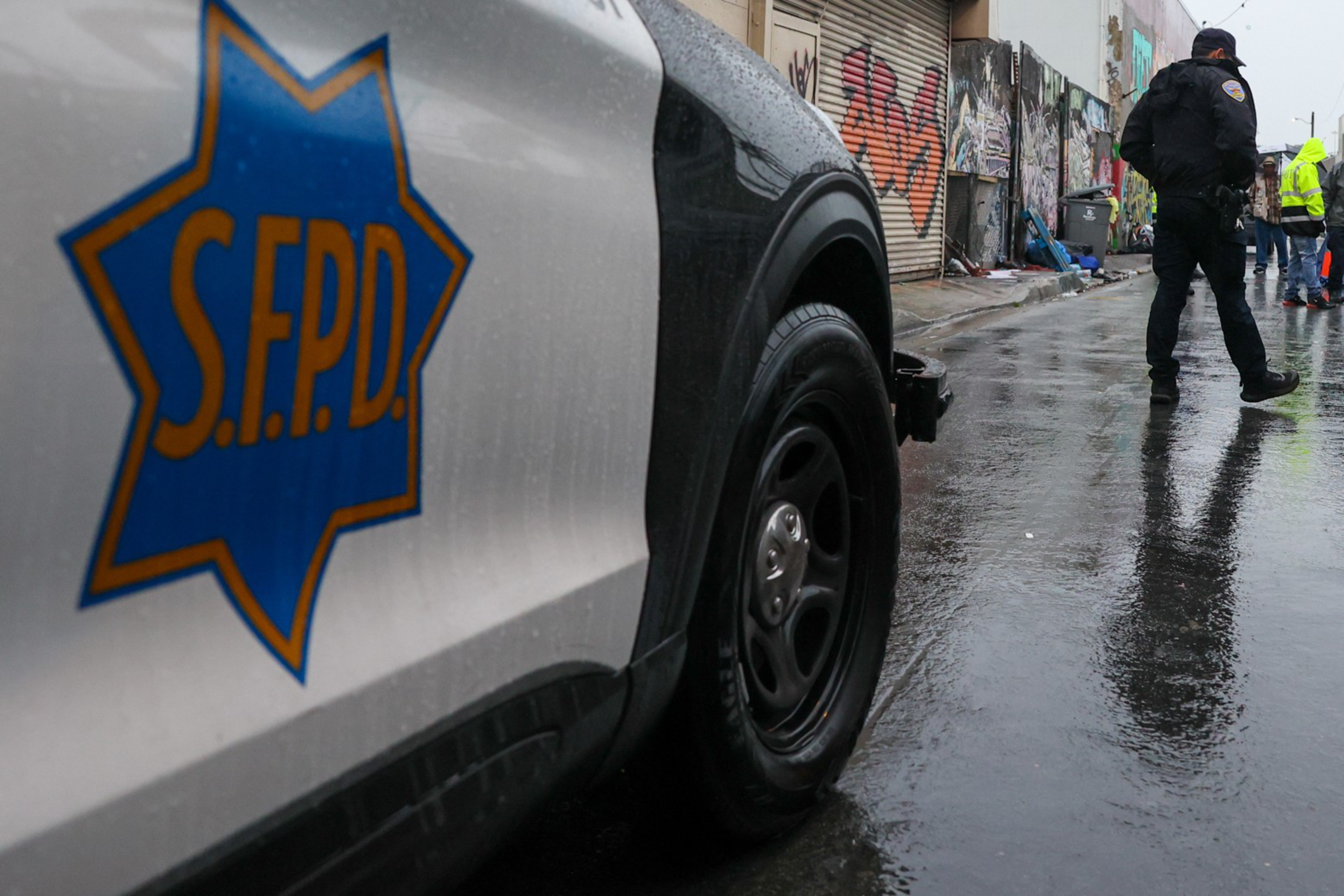 A rainy street with an S.F.P.D. police car in the foreground and officers standing near graffiti-covered walls in the background.