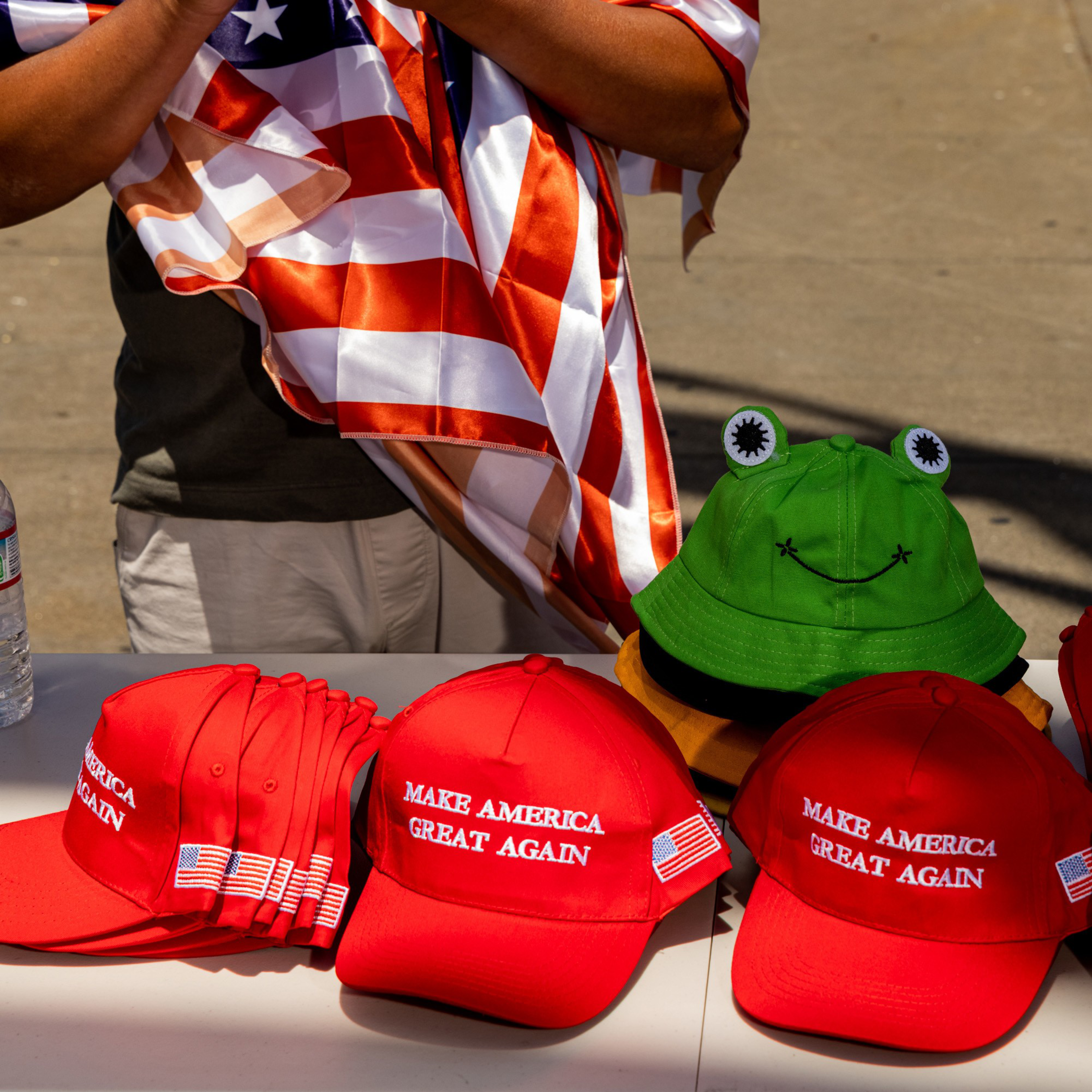 Red &quot;Make America Great Again&quot; hats are displayed on a table alongside a green hat with frog eyes and a smiley face. An individual holds an American flag.
