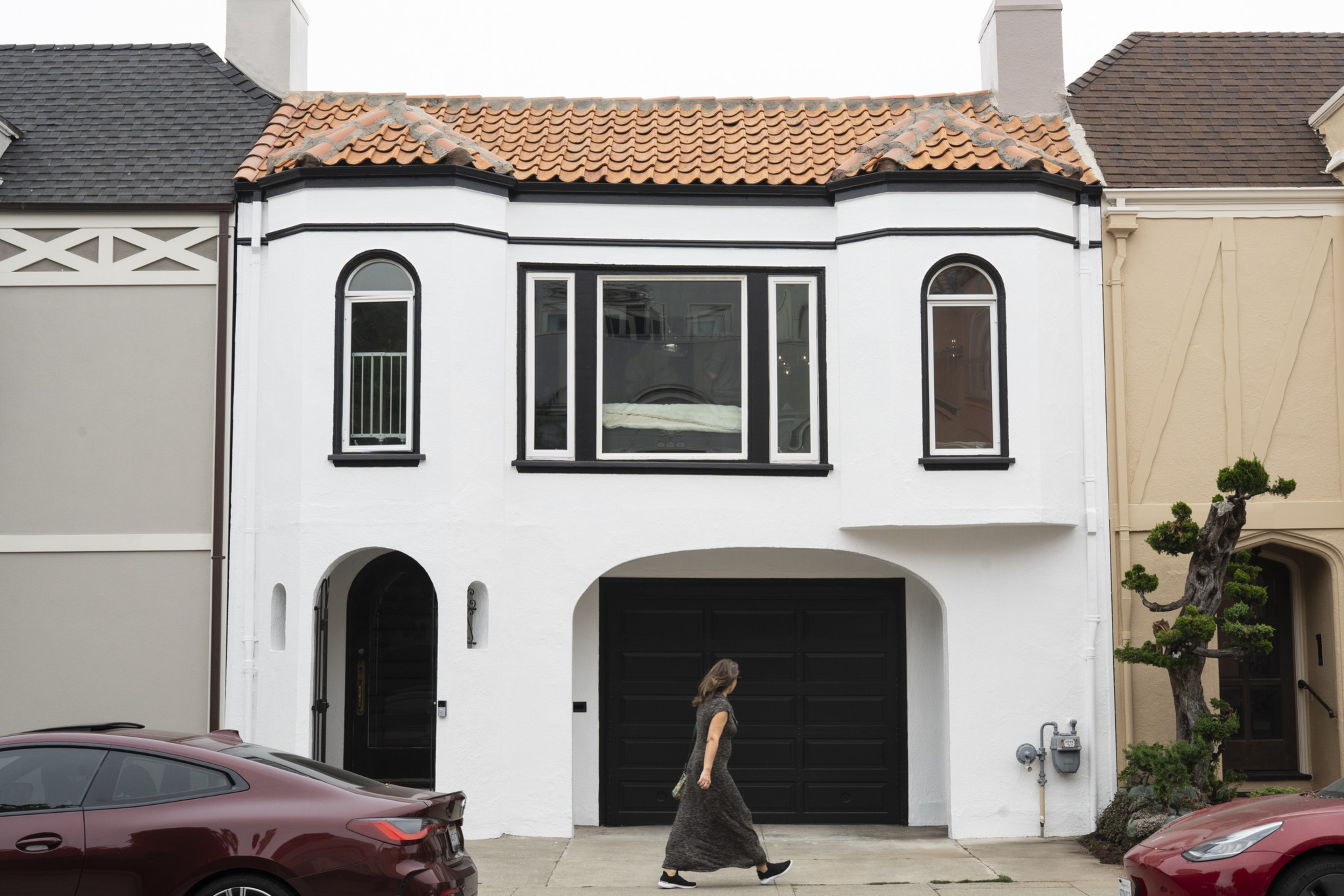 A white two-story house with black trim and a terracotta tiled roof sits between two other houses, with a black garage door and a woman walking past.